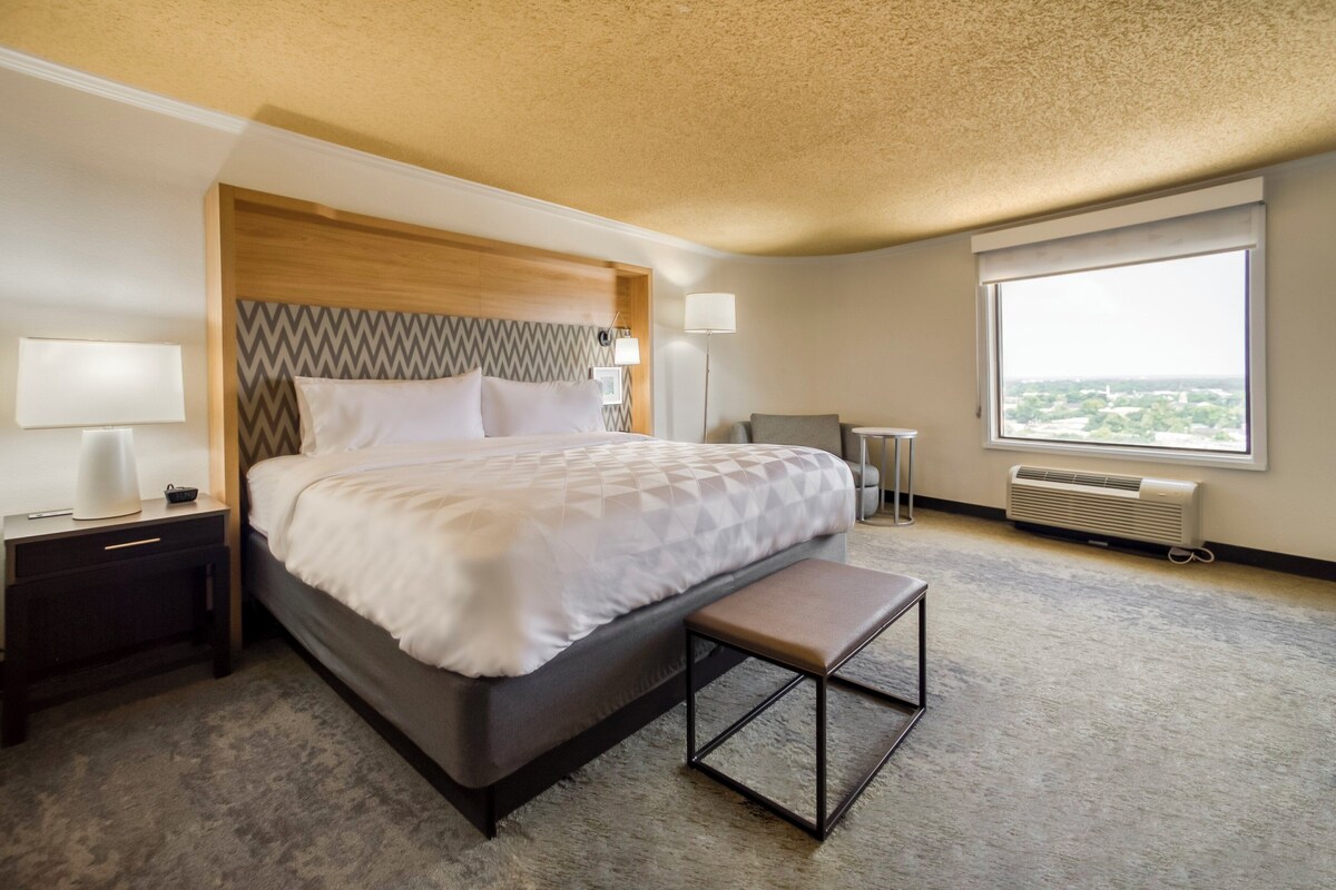 A hotel room with a large bed, bedside lamps, a bench, a desk, and a window with a view.