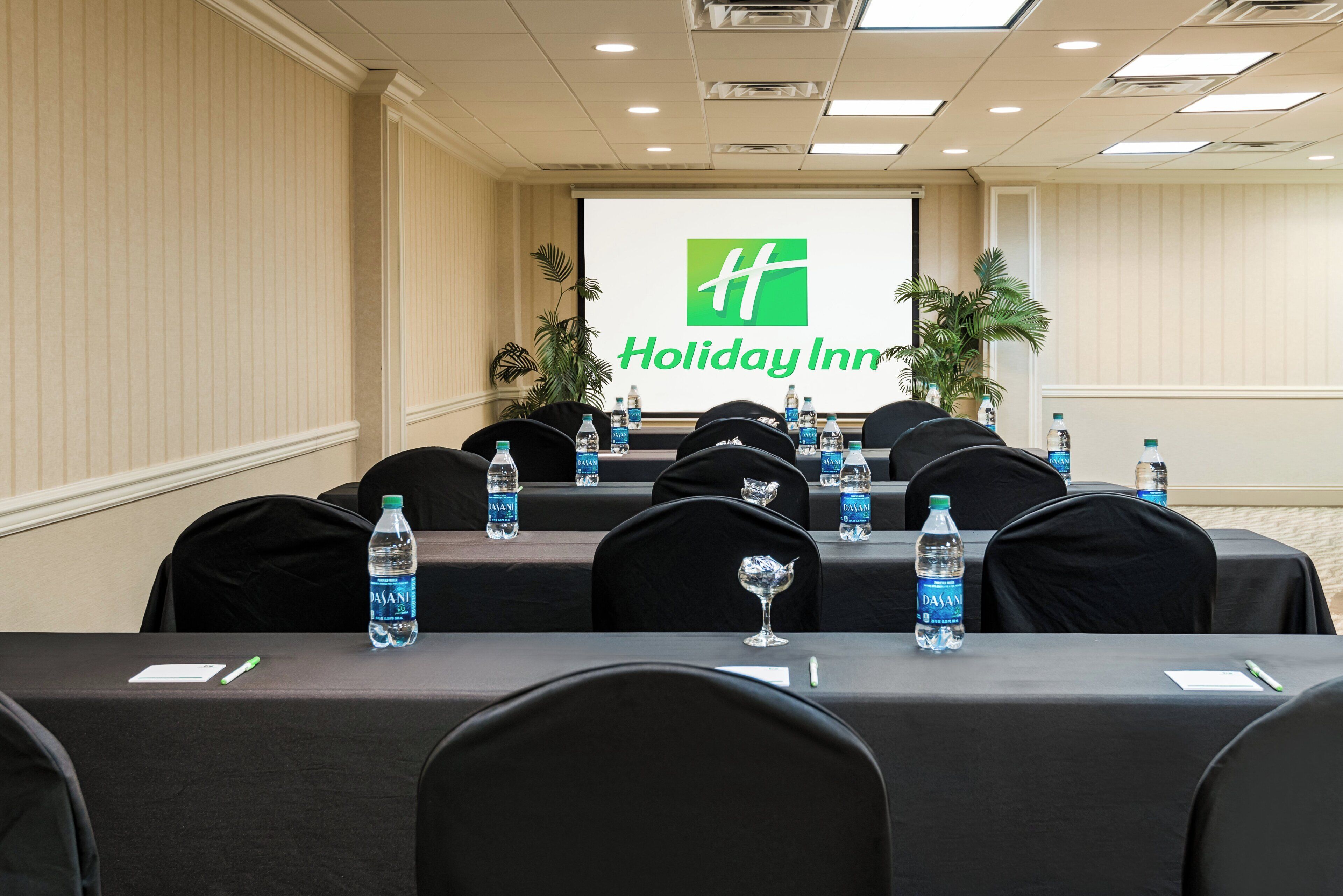 A conference room with a Holiday Inn logo on the screen, rows of tables with water bottles, and black chairs.