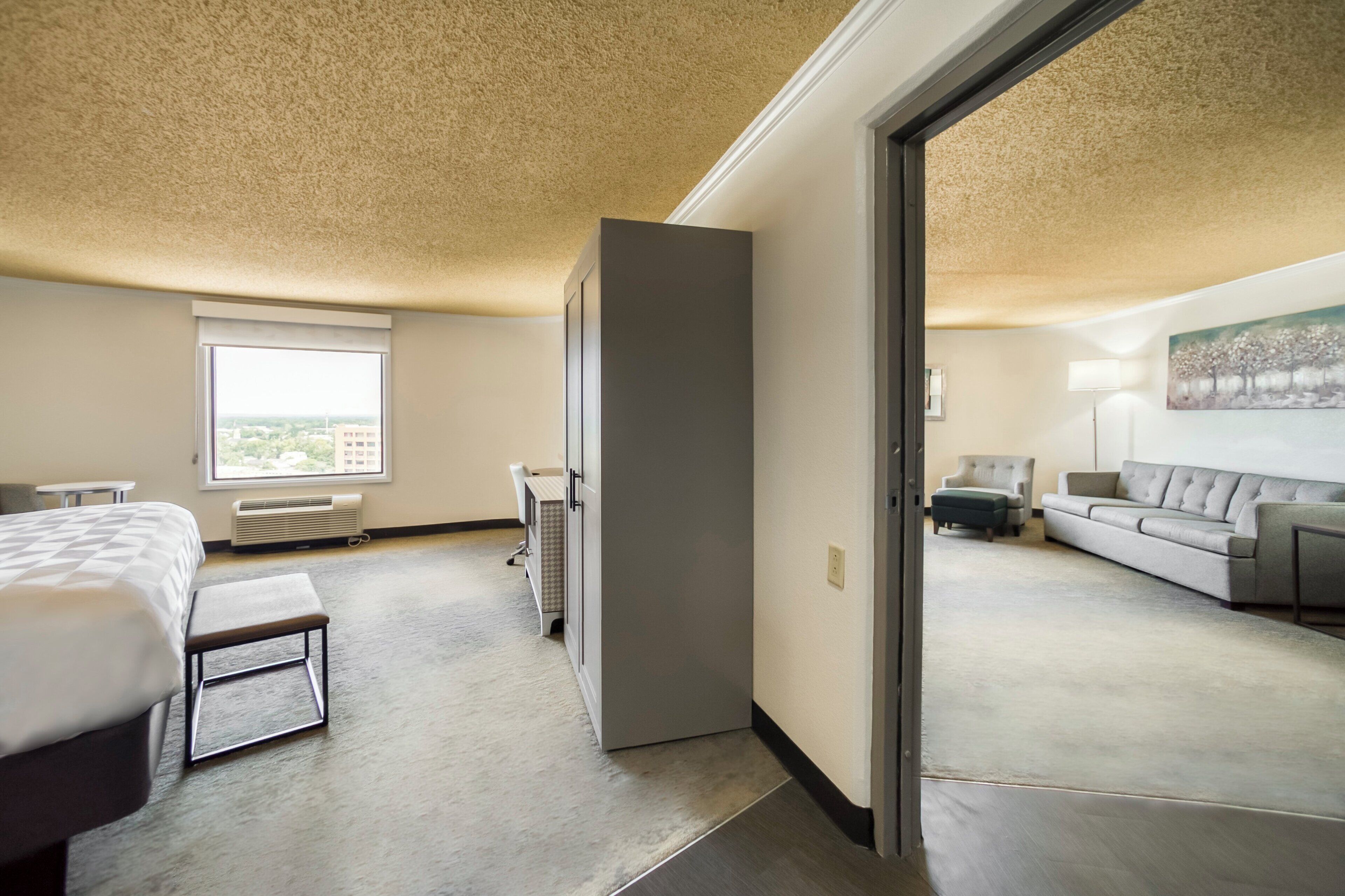 A hotel room with a bed, a desk, a chair, a sofa, a painting, and a view of the city through a window.