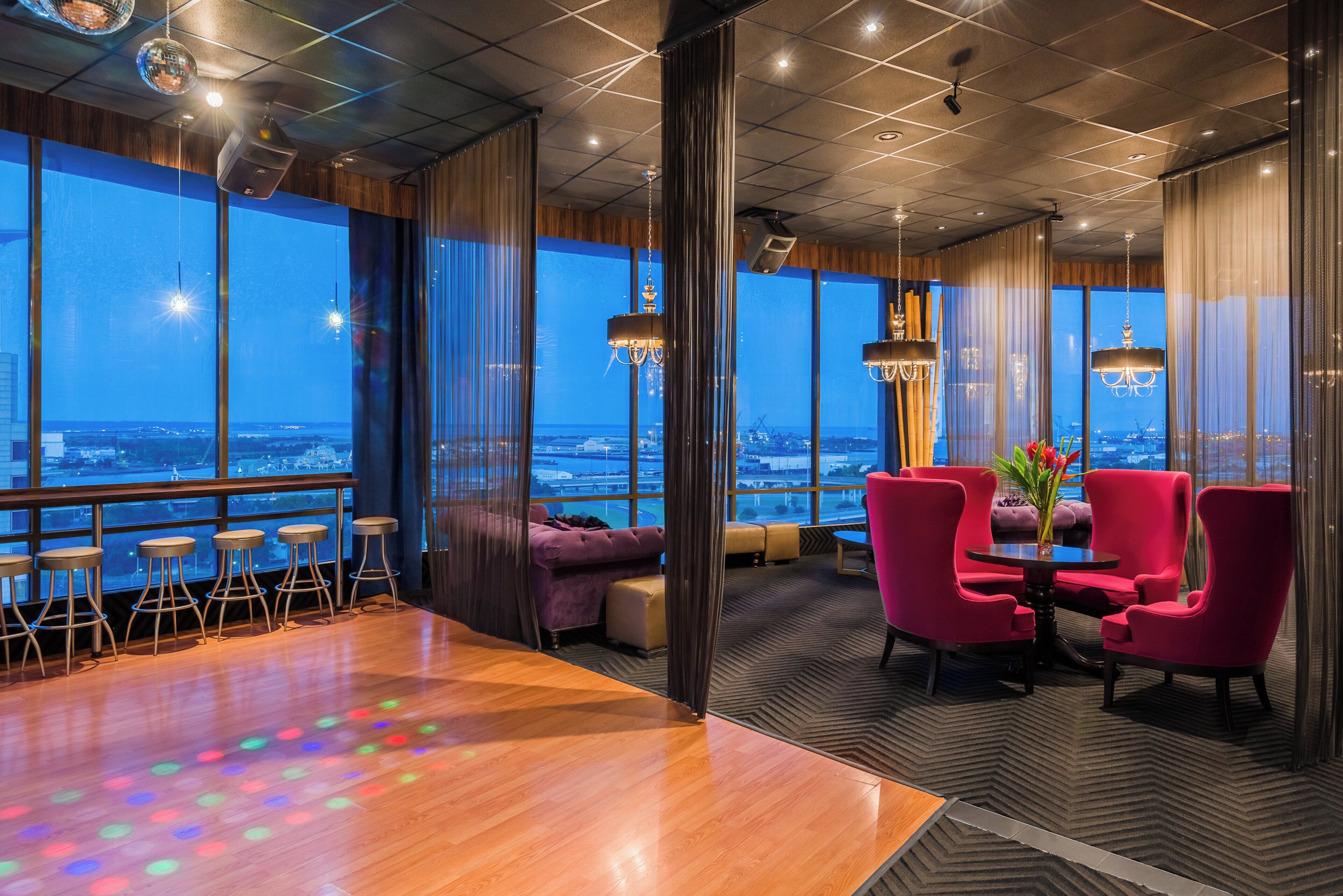 Uma área de estar moderna com vista para a paisagem urbana, incluindo um bar com banquetas, uma área de assentos com cadeiras vermelhas e uma grande janela com vista panorâmica.