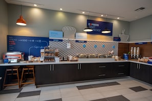 A modern kitchen area with a breakfast bar, counter with food items, and a sign indicating breakfast options. - Holiday Inn Mobile-Dwtn/Hist. District by IHG (Mobile)