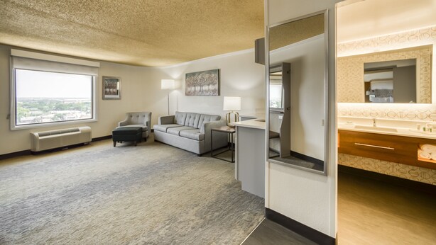 A hotel room with a large window, a gray sofa, a small table, a lamp, and a bathroom with a mirror and sink. - Holiday Inn Mobile-Dwtn/Hist. District by IHG (Mobile)