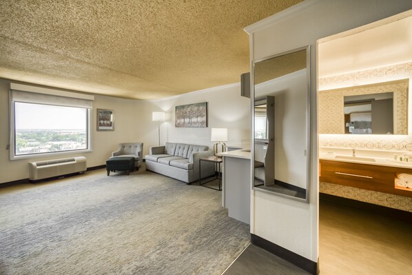 A hotel room with a large window, a gray sofa, a small table, a lamp, and a bathroom with a mirror and sink. - Holiday Inn Mobile-Dwtn/Hist. District by IHG (Mobile)