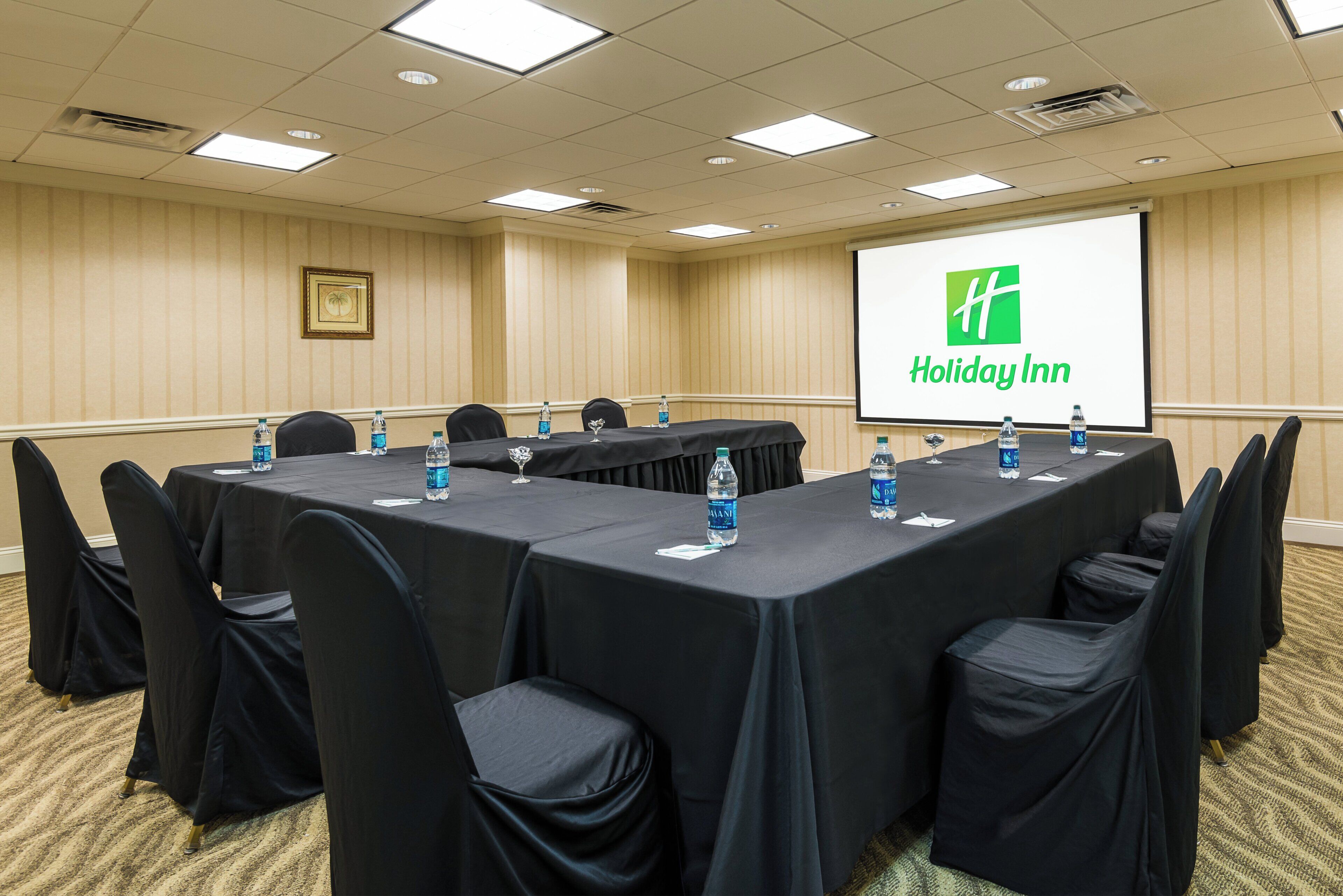 Un salón de conferencias con una larga mesa, sillas, botellas de agua y una pantalla que muestra el logotipo de Holiday Inn.