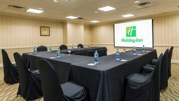 Un salón de conferencias con una larga mesa, sillas, botellas de agua y una pantalla que muestra el logotipo de Holiday Inn.