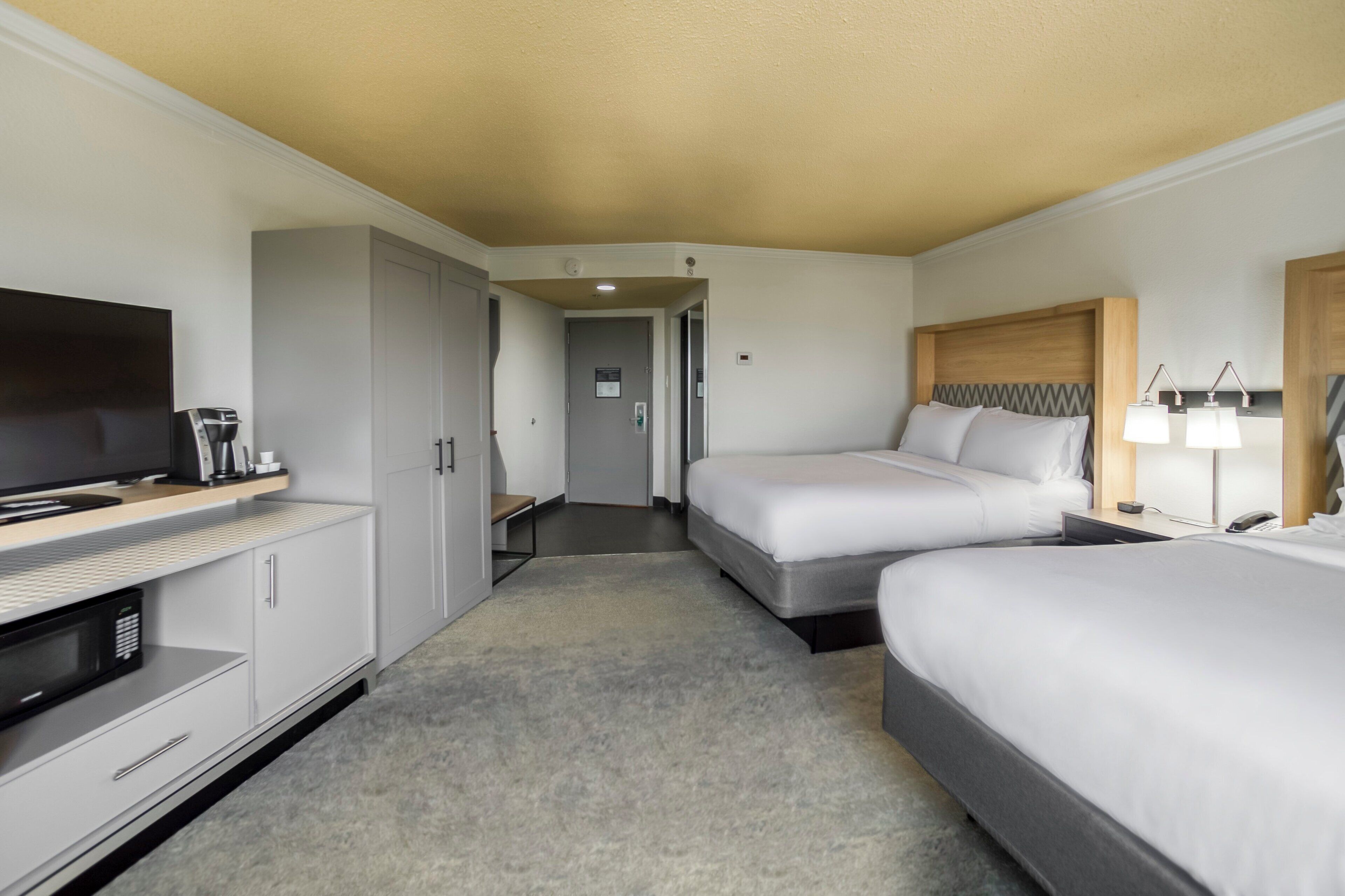 A hotel room with a TV, a desk with a coffee maker, two beds, a nightstand with lamps, and a door leading to a hallway.