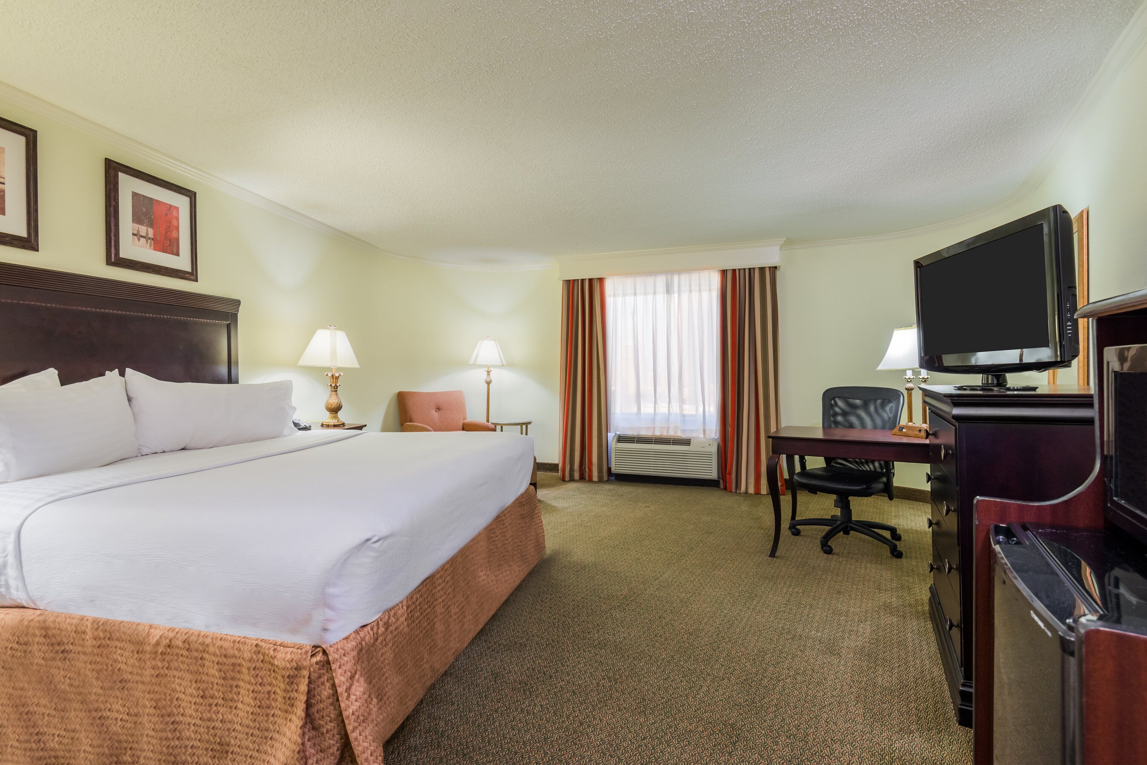 A hotel room with a large bed, a desk with a television, a chair, and an air conditioning unit.