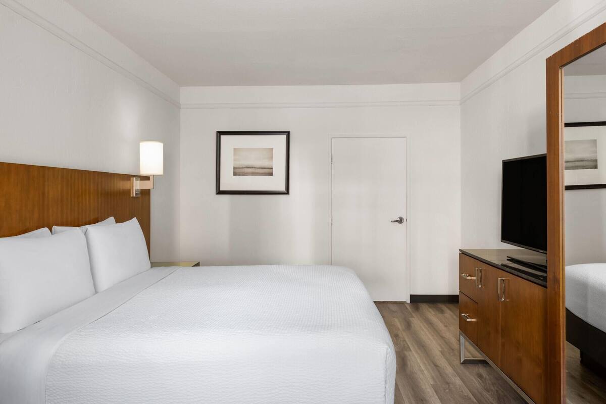 Een hotelkamer met een groot bed, een houten hoofdeinde, een nachtkastje met een lamp, een televisie en een ingelijst schilderij aan de muur.
