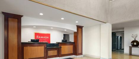 Un área de recepción con un mostrador de madera, un cartel que dice "RAMADA SUITES BY WYNDHAM" y un felpudo negro con la palabra "RAMADA".