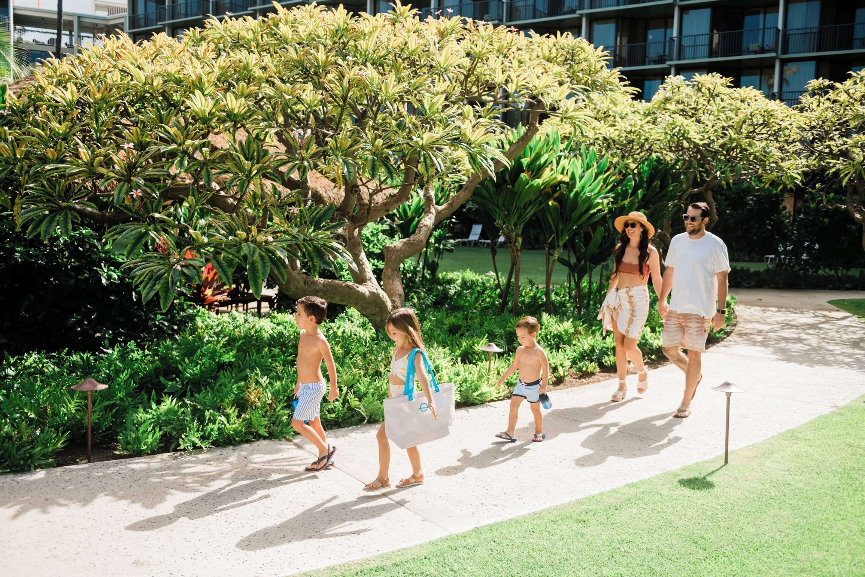 A pathway with a group of people walking, surrounded by greenery and a building in the background.