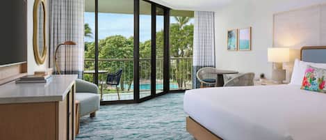 A hotel room with a bed, a desk, a chair, a balcony with a view of trees, and a ceiling fan.