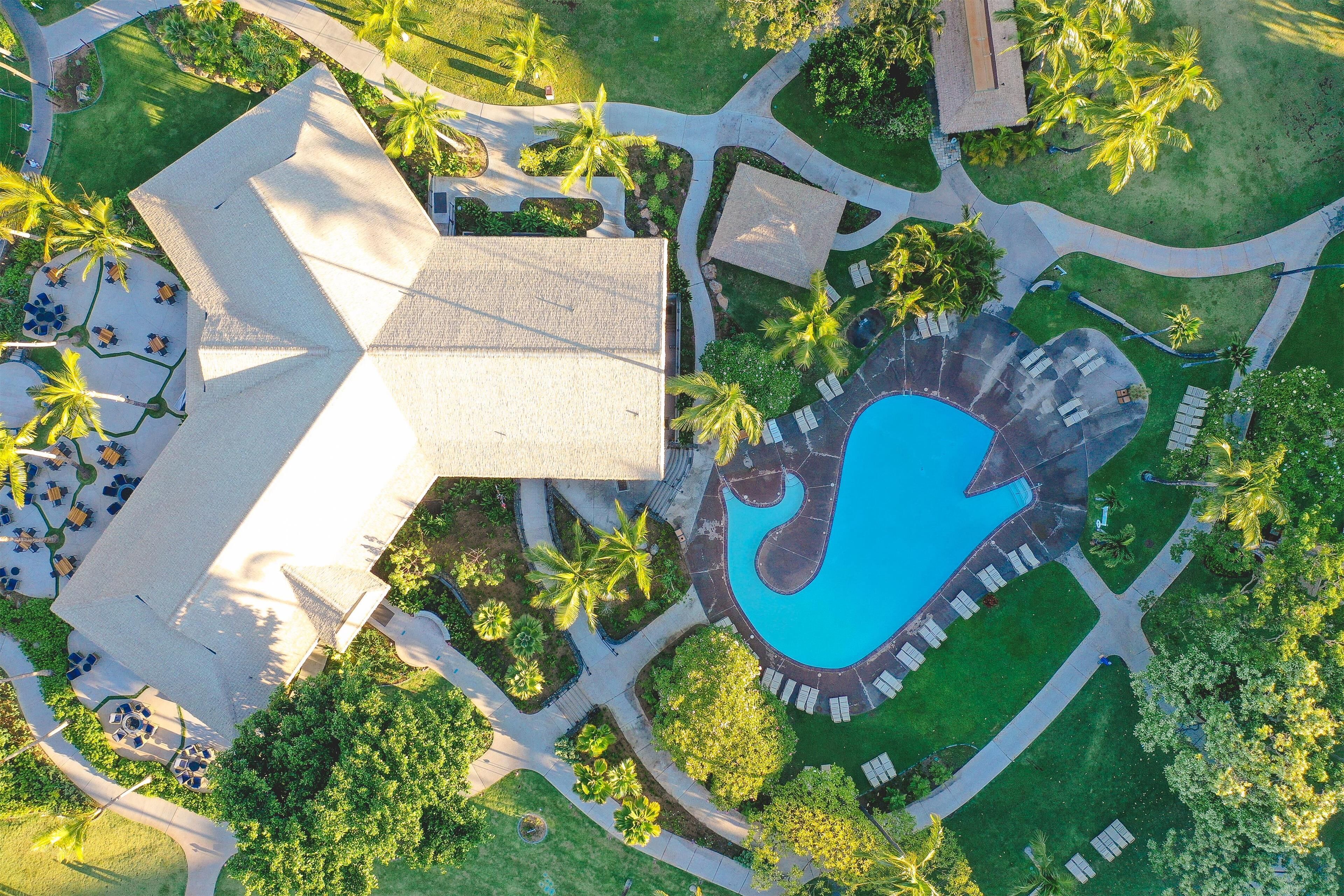 Eine Vogelperspektive zeigt ein Resort mit Schwimmbad, Palmen und einem großen Gebäude mit markanter Dachkonstruktion.