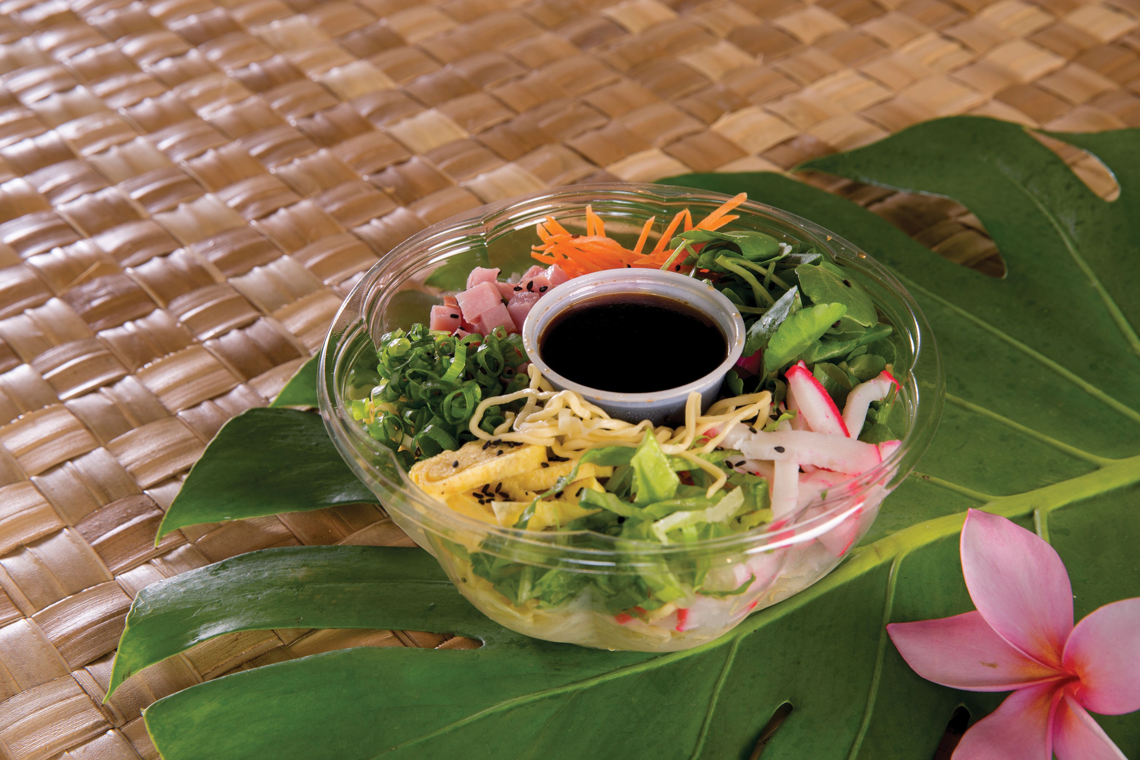 Uma saladeira transparente com salada e diversos vegetais, acompanhada de uma pequena tigela de molho sobre um tapete trançado.