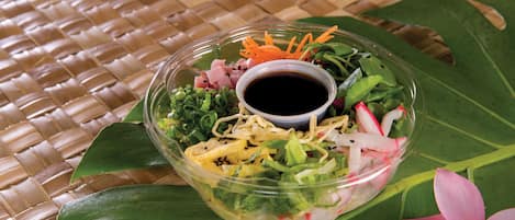 A clear bowl of salad with various vegetables and a small cup of sauce on a woven mat.