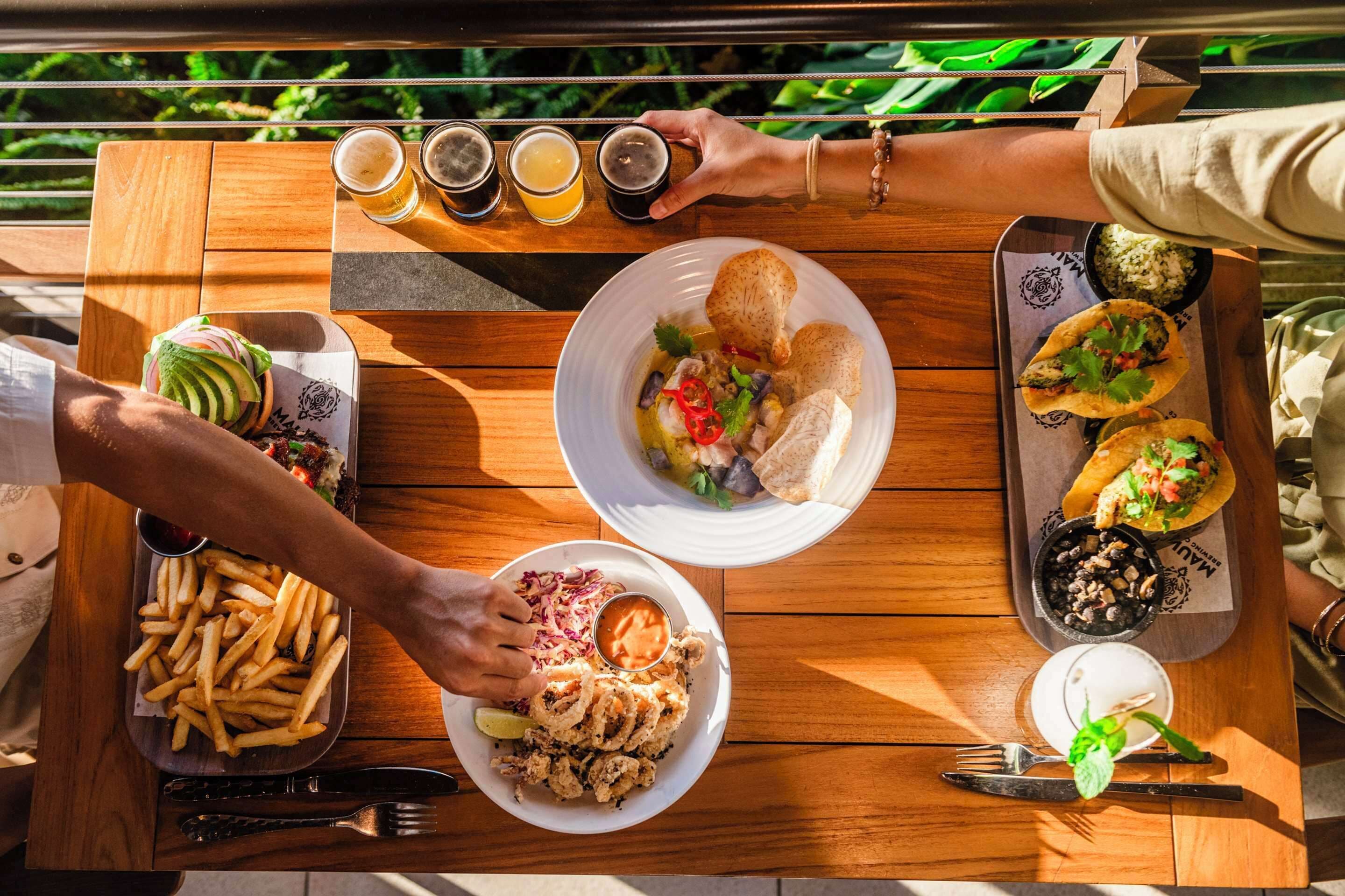 Uma mesa de madeira com vários pratos, incluindo um hambúrguer, tacos e uma salada.