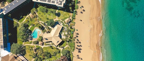 An aerial view of a resort with a beach, a swimming pool, and a large building with solar panels.