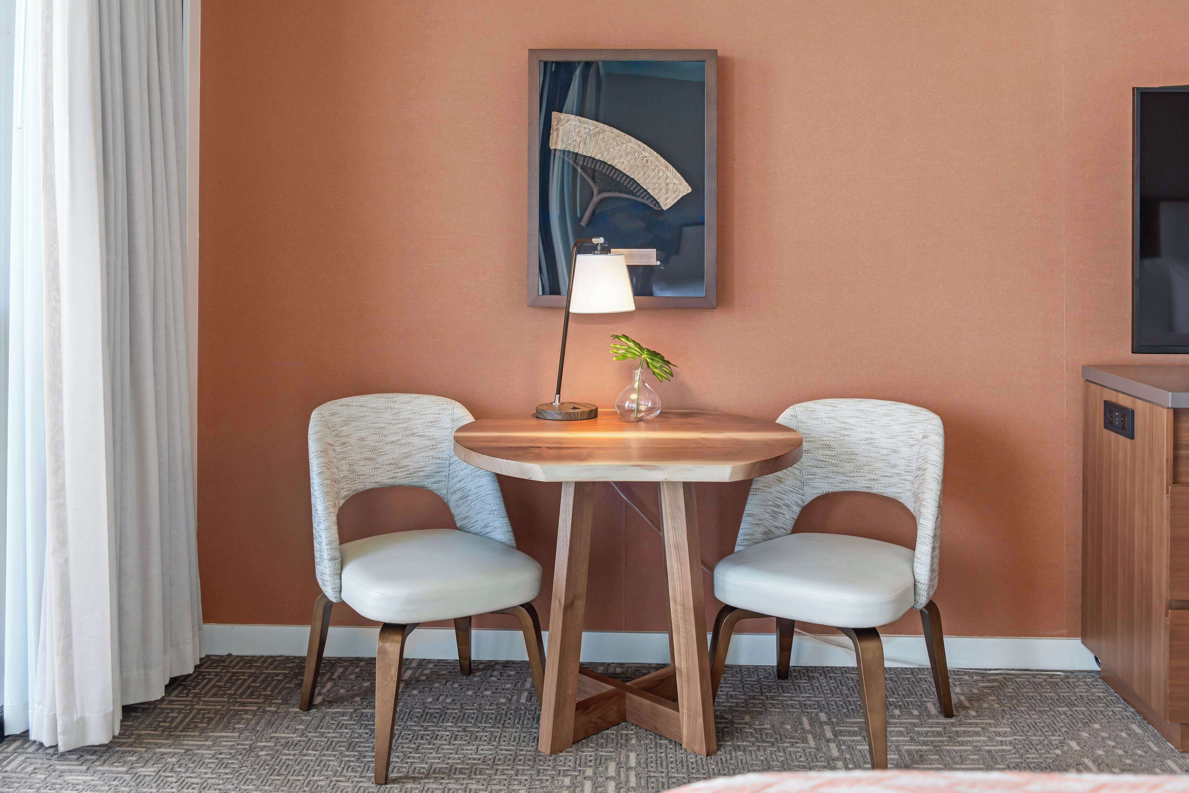 A hotel room with a round wooden table, two upholstered chairs, a floor lamp, a small potted plant, and a framed artwork on the wall.