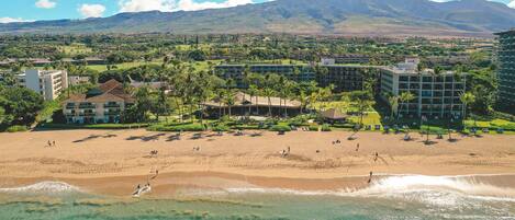 A beach resort with multiple buildings, a sandy beach, and a mountain in the background.
