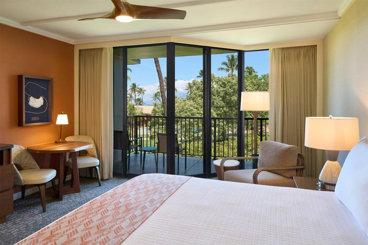 Una camera d'albergo con un letto grande, una scrivania, due sedie, un balcone con vista su palme e l'oceano, e una ventola a soffitto.