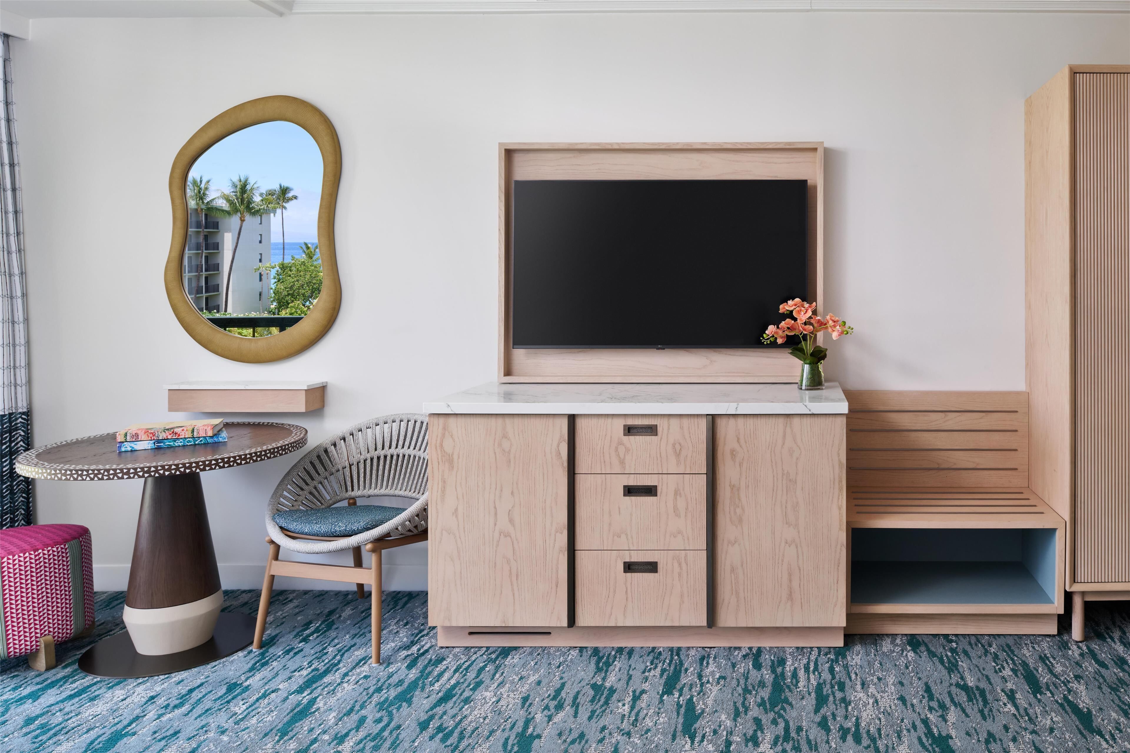 Une pièce comprenant un téléviseur, un meuble en bois, une table ronde, une chaise en osier et un miroir qui reflète un bâtiment.
