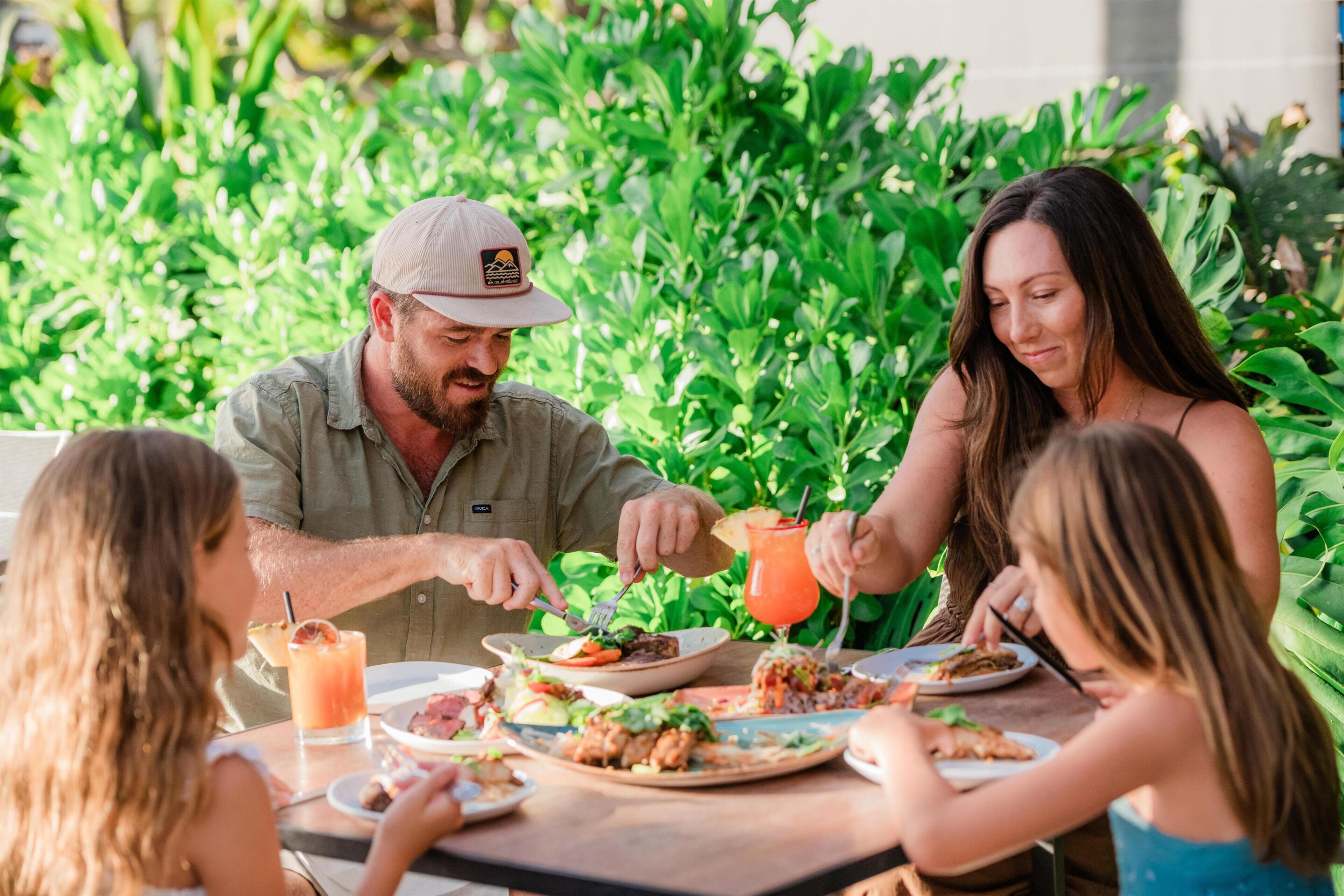 Une famille prend son repas à l'extérieur, avec en arrière-plan une végétation luxuriante et verdoyante.