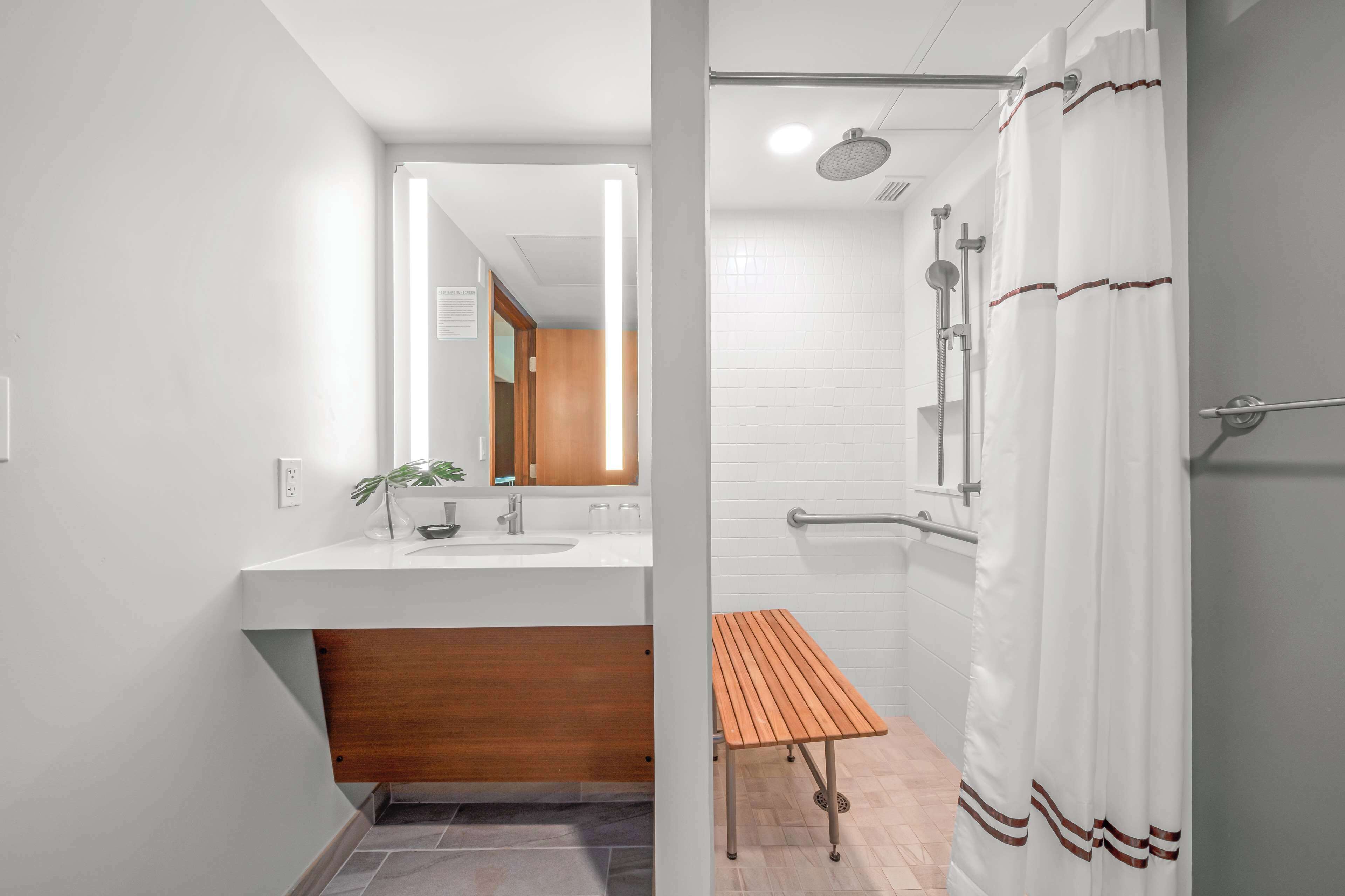 A modern bathroom with a white sink, wooden vanity, a bench, a shower with a curtain, and a mirror.