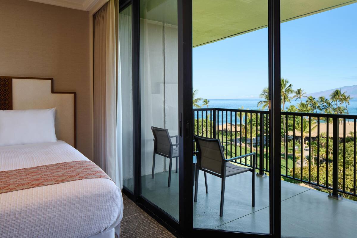 Una camera d'hotel con un letto, una porta scorrevole in vetro che dà su un balcone con sedie e vista sulla spiaggia.