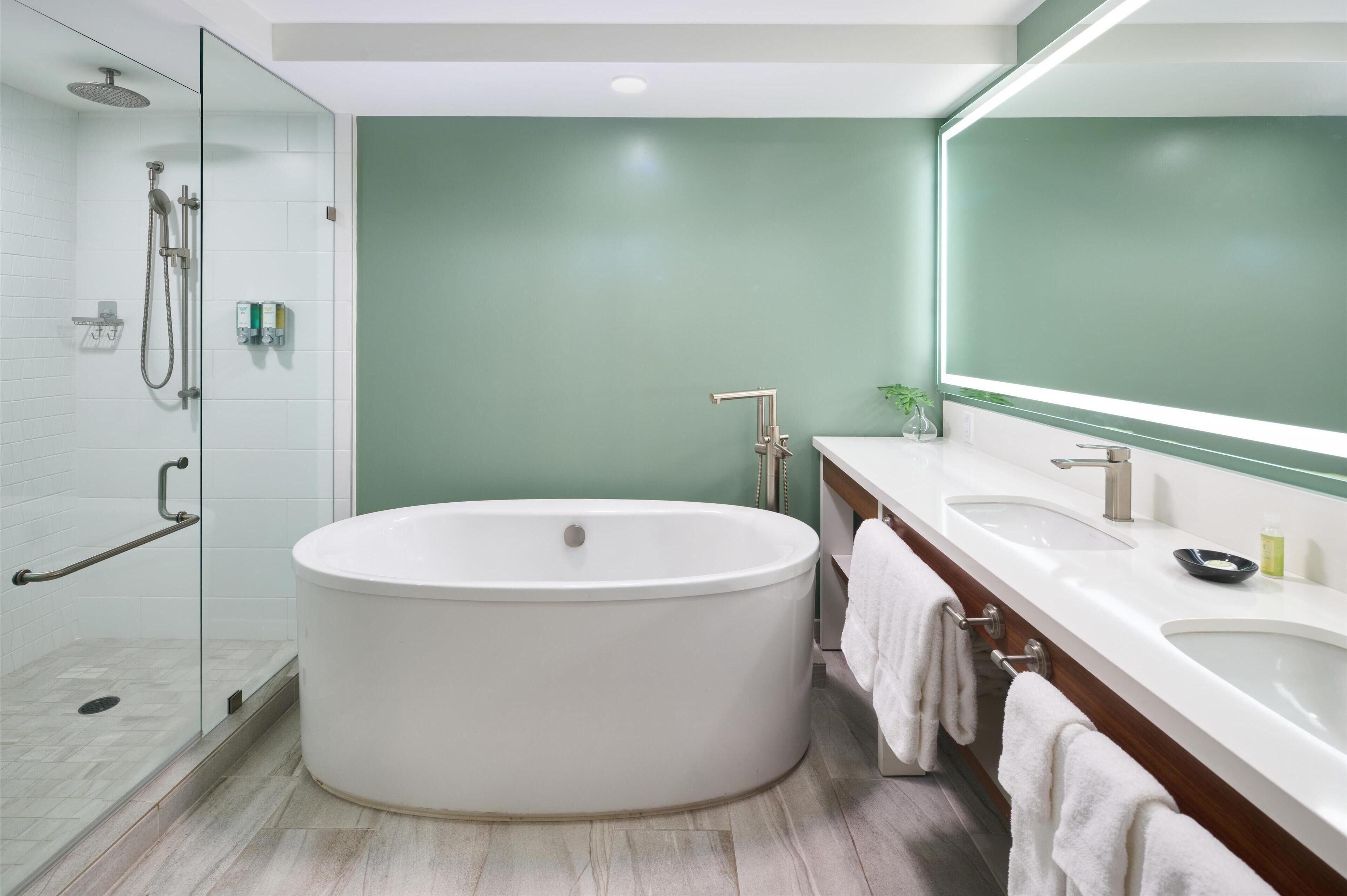 Ein modernes Badezimmer mit einer großen Badewanne, zwei Waschbecken, einer Duschkabine aus Glas und einem Spiegel mit Lichtleiste.