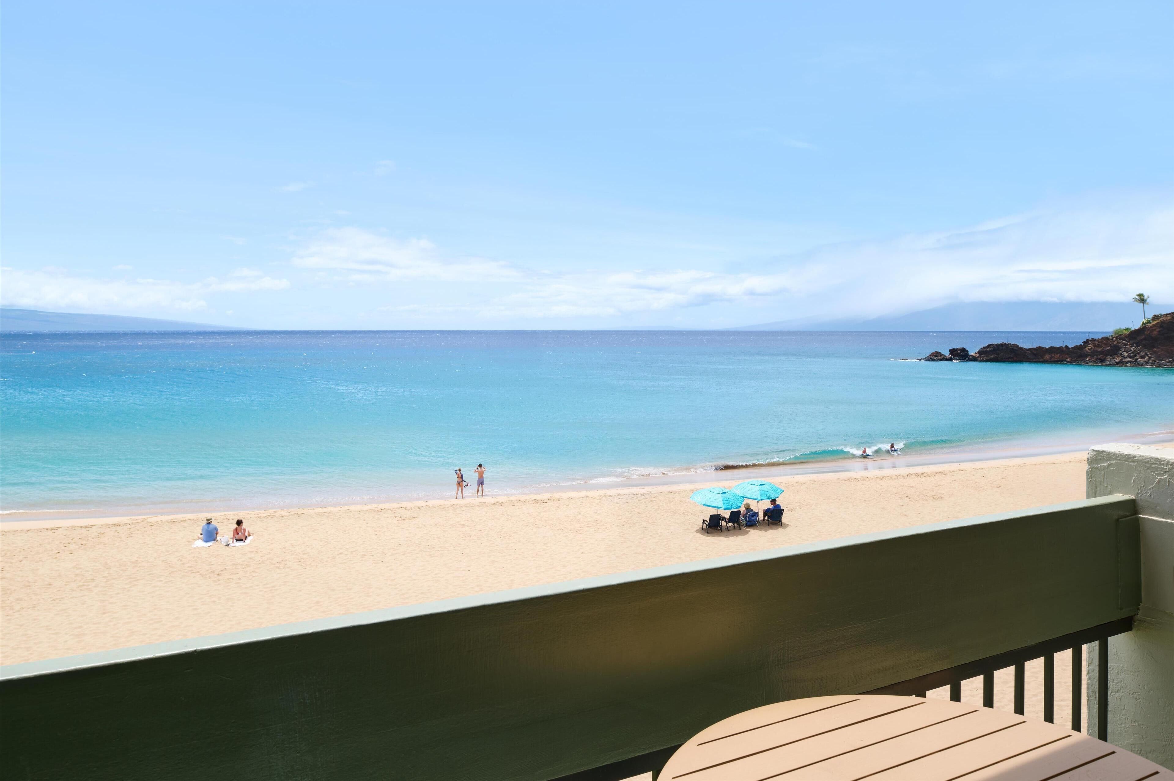 Blick auf einen Strand vom Balkon aus, mit blauem Himmel, Sandstrand und einigen Personen und Sonnenschirmen.