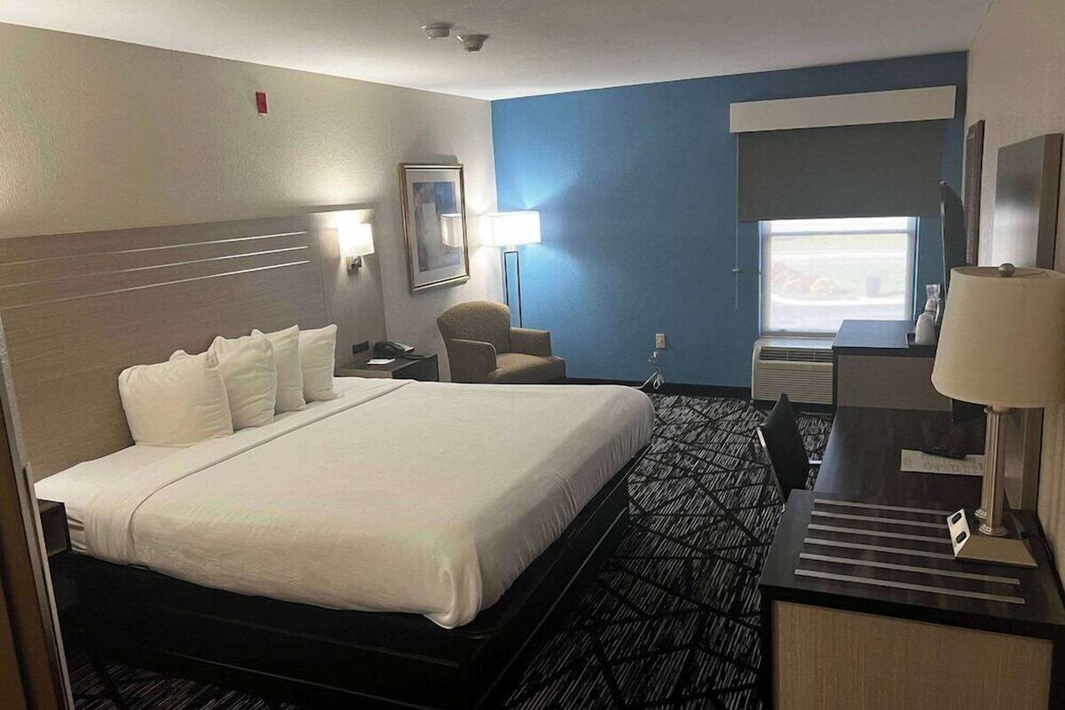 Quarto de hotel com uma cama grande, uma escrivaninha com luminária, uma cadeira, uma janela e uma televisão fixada na parede.