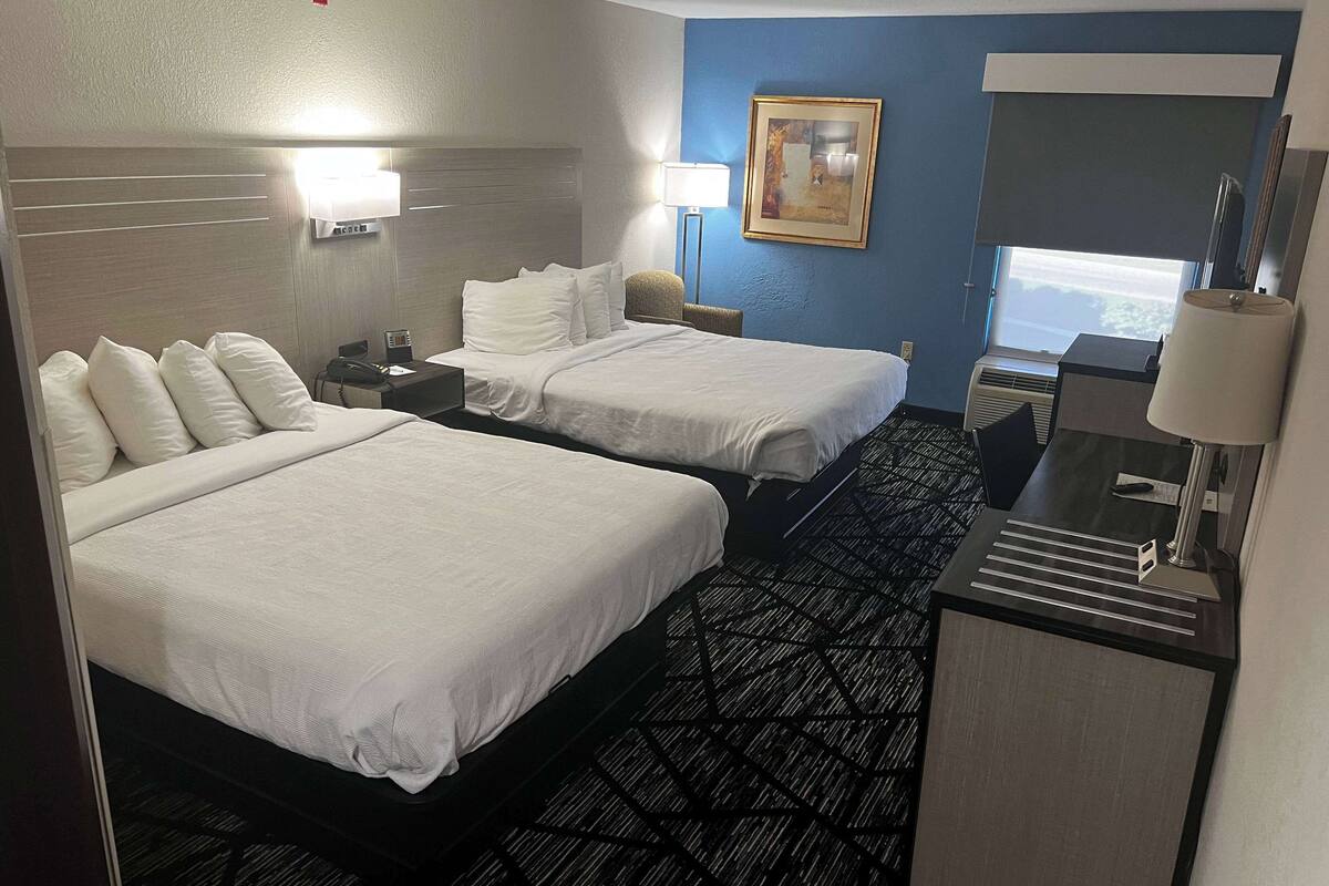Quarto de hotel com duas camas, uma escrivaninha, uma lâmpada, um quadro e uma janela.