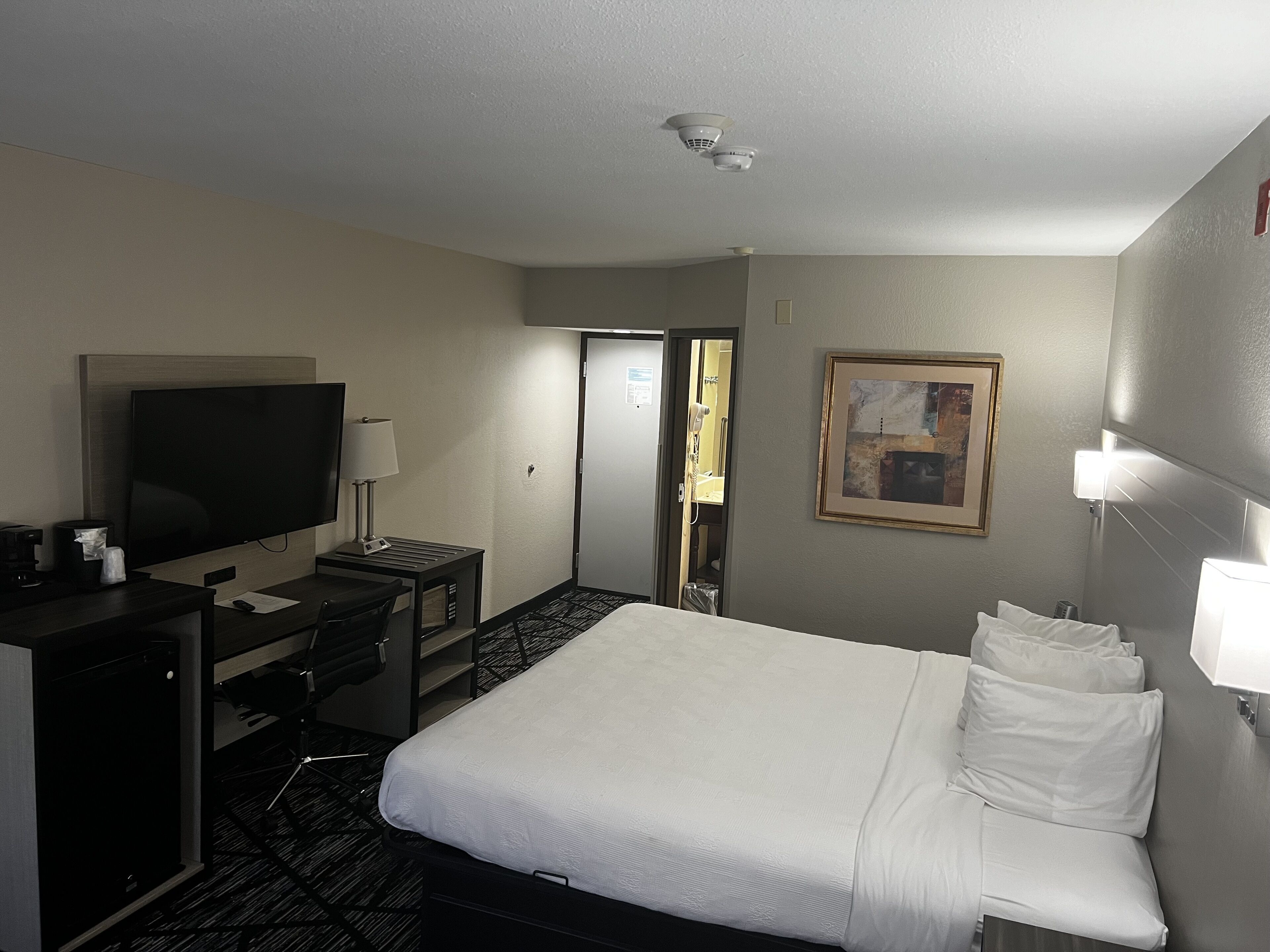 A hotel room with a bed, a desk with a TV, a lamp, and a painting on the wall.