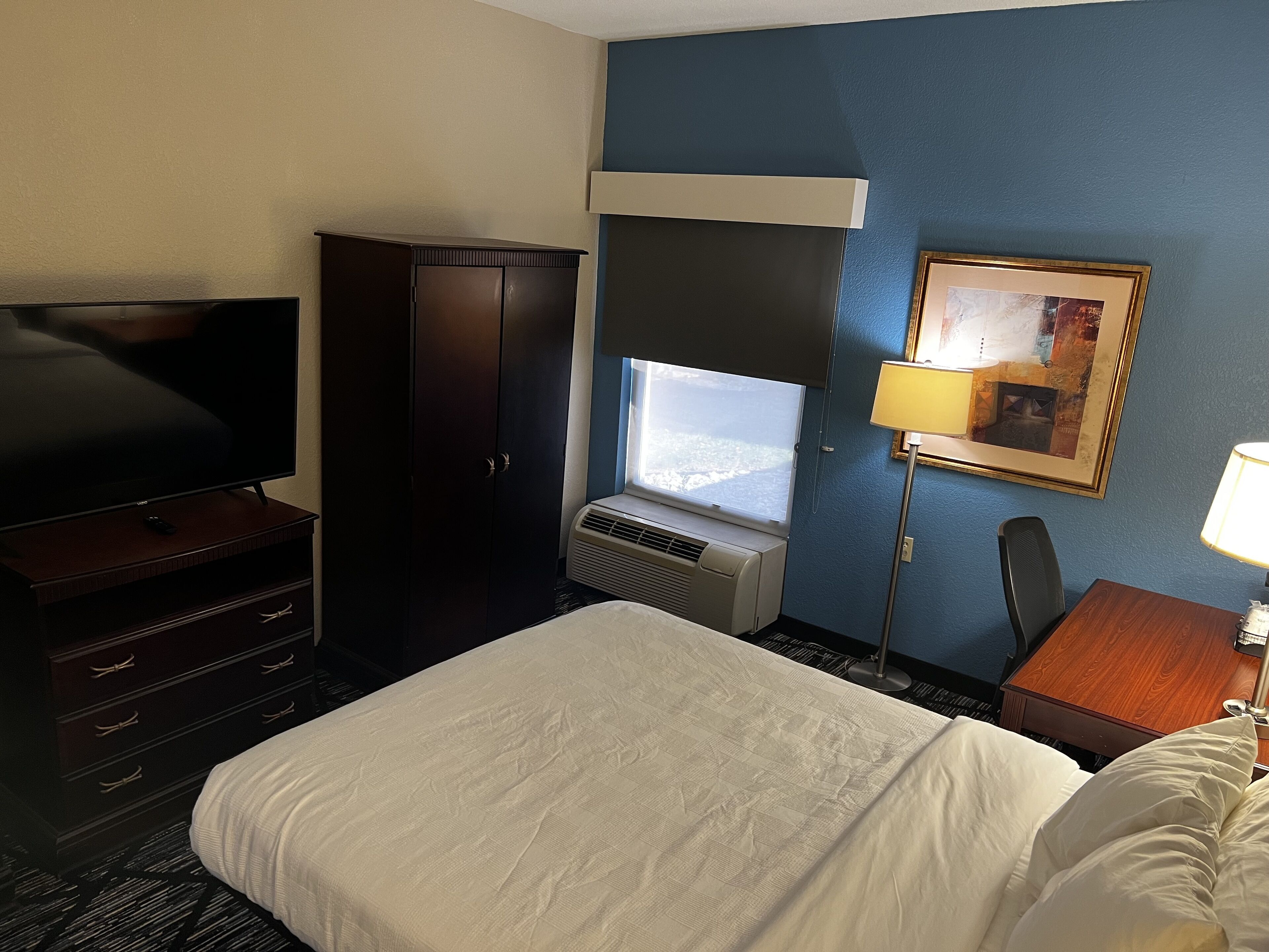 A hotel room with a bed, a TV, a wardrobe, a desk with a lamp, and a window with a view.