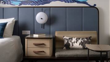A hotel room with a bed, bedside table, nightstand, and a small seating area with a pillow.