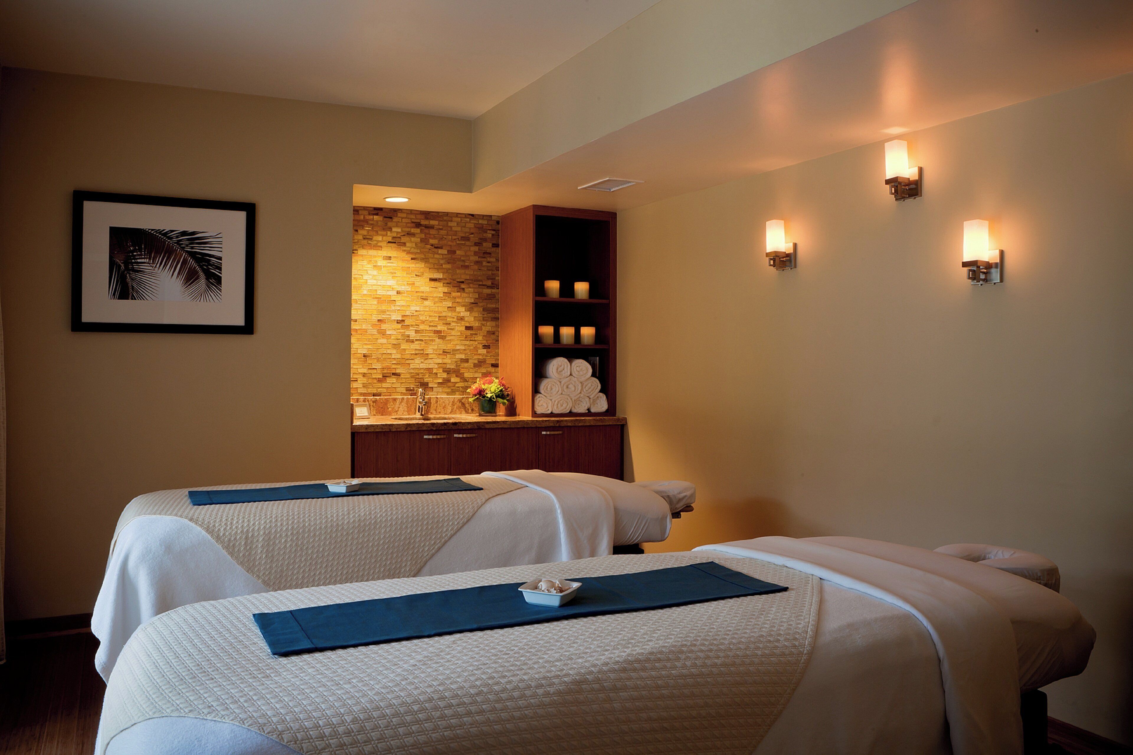 A spa room with two massage tables, a stone wall, a wooden cabinet with towels, and wall-mounted lights.
