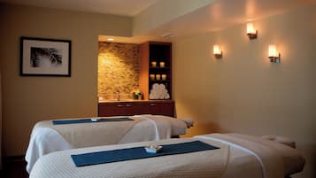 A spa room with two massage tables, a stone wall, a wooden cabinet with towels, and wall-mounted lights.