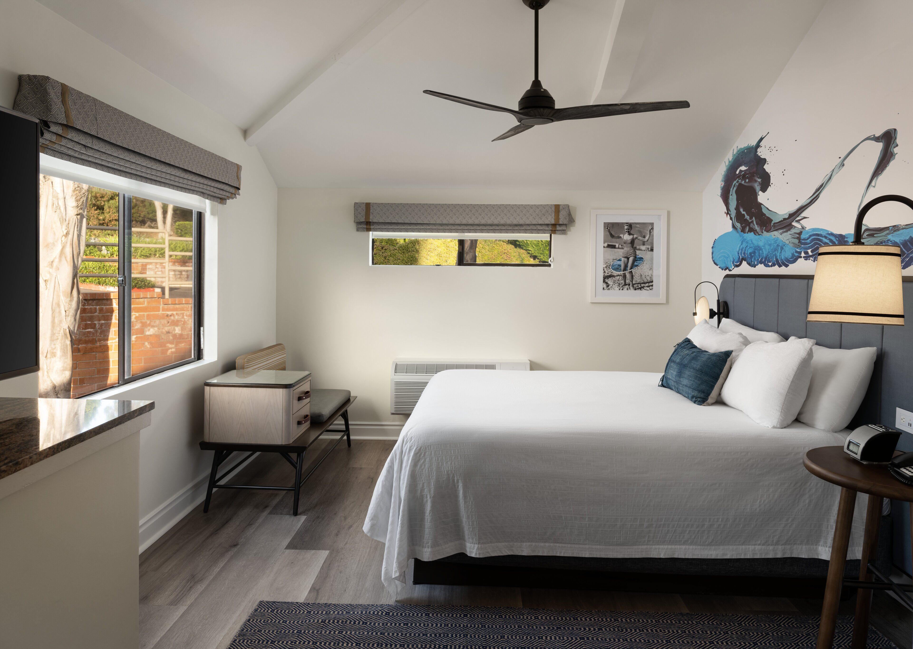 A bedroom with a bed, bedside table, lamp, and a view of the outdoors through a window.