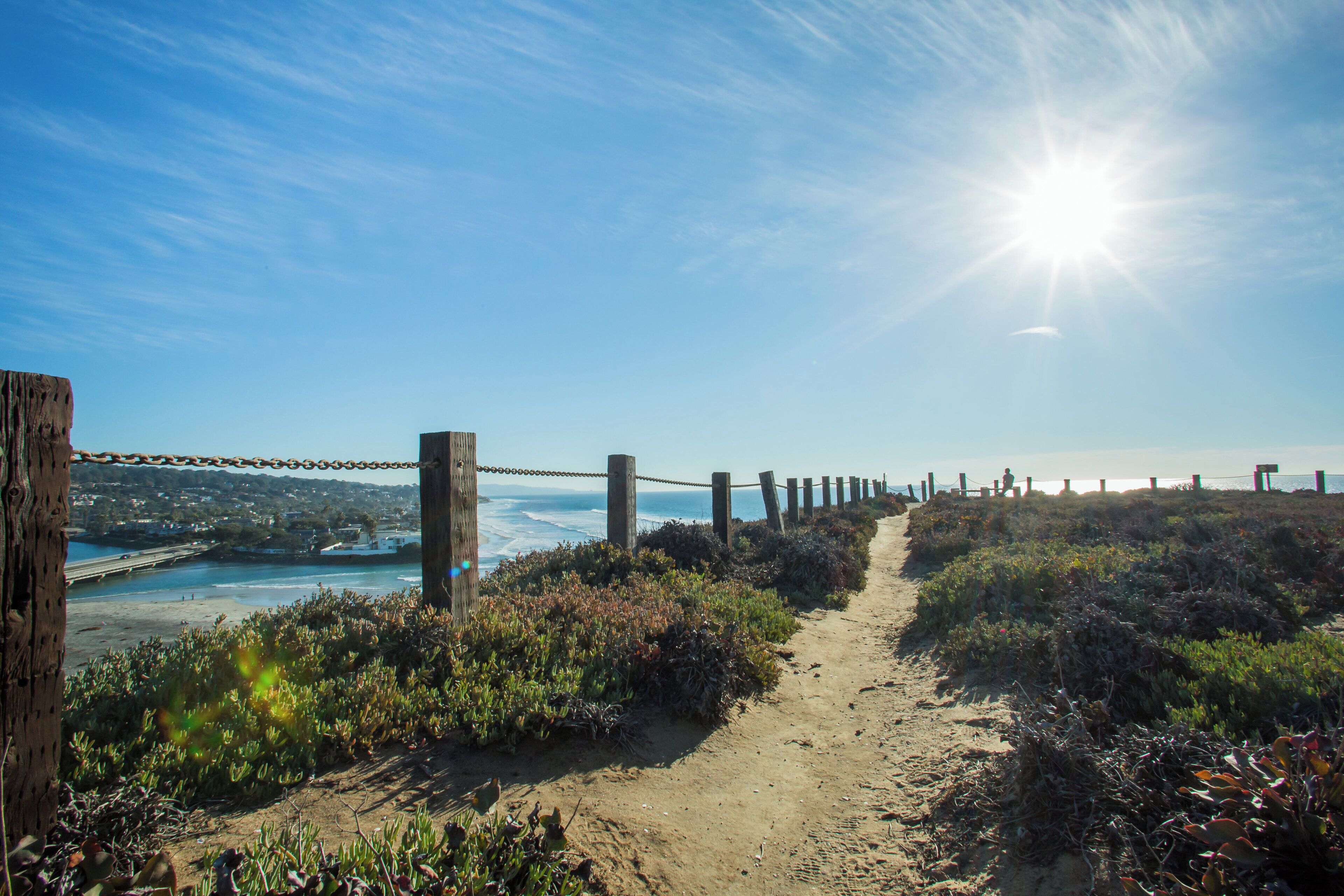 A coastal path with a chain-link fence, leading to a beach and ocean view.