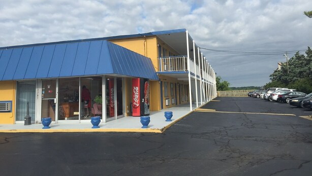 Ein Motel mit blauem Dach und gelben Wänden, mit Coca-Cola Schildern, einem Parkplatz und blauem Himmel.