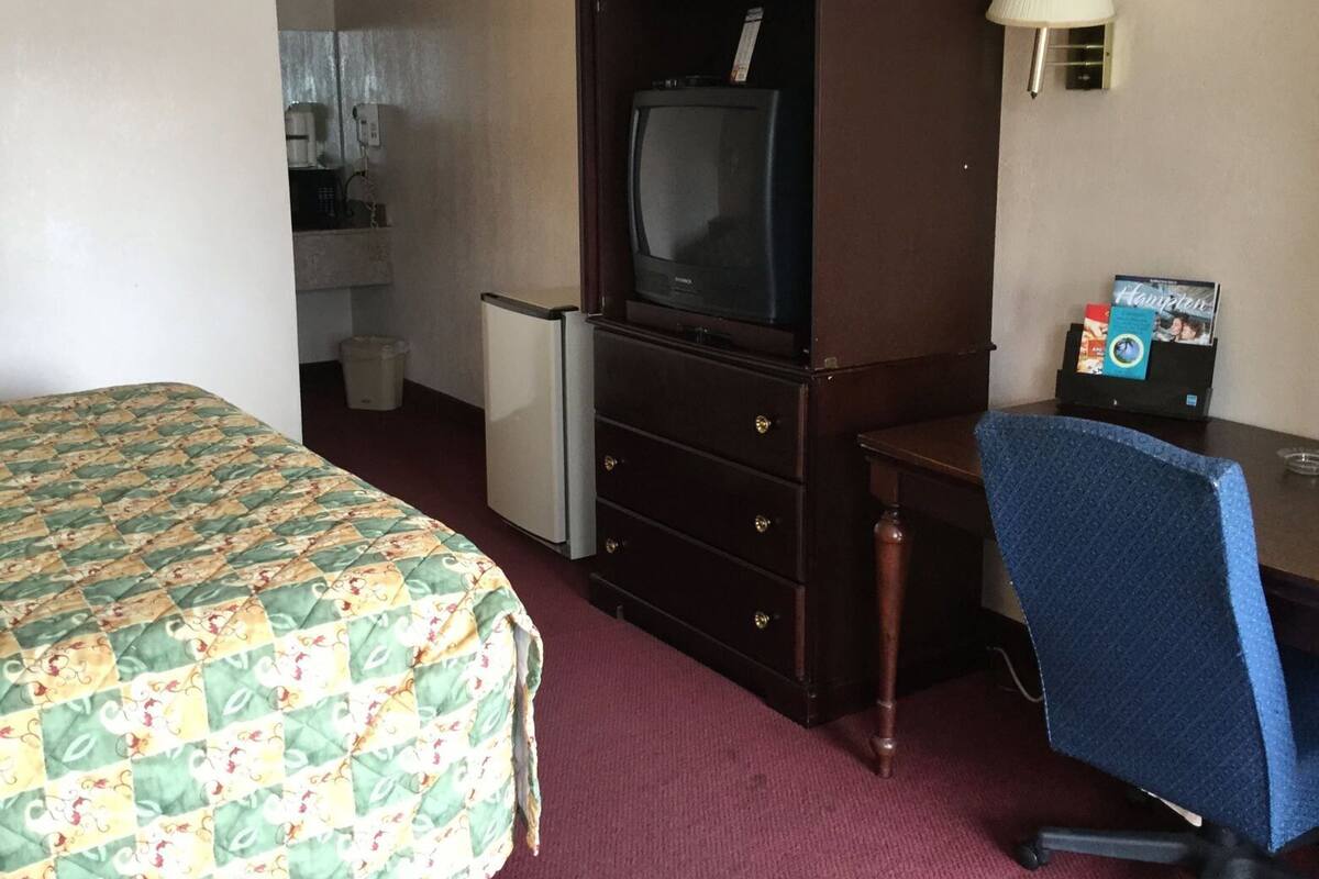 A hotel room with a bed, a desk with a chair, a television, a refrigerator, and a lamp.