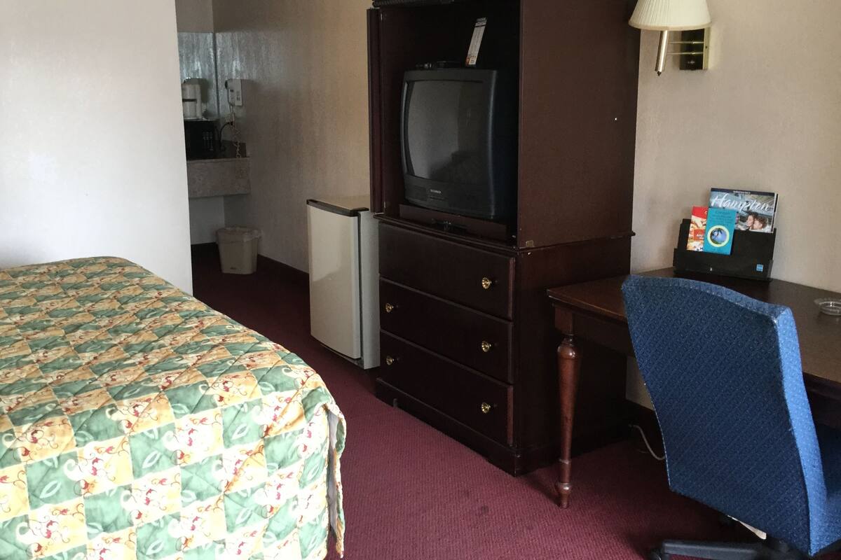 A hotel room with a bed, a desk with a chair, a television, a refrigerator, and a lamp.