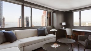 A modern living room with a sofa, two round tables, and a desk with a chair. Large windows offer a view of the city skyline. - New York Marriott Downtown (New York)