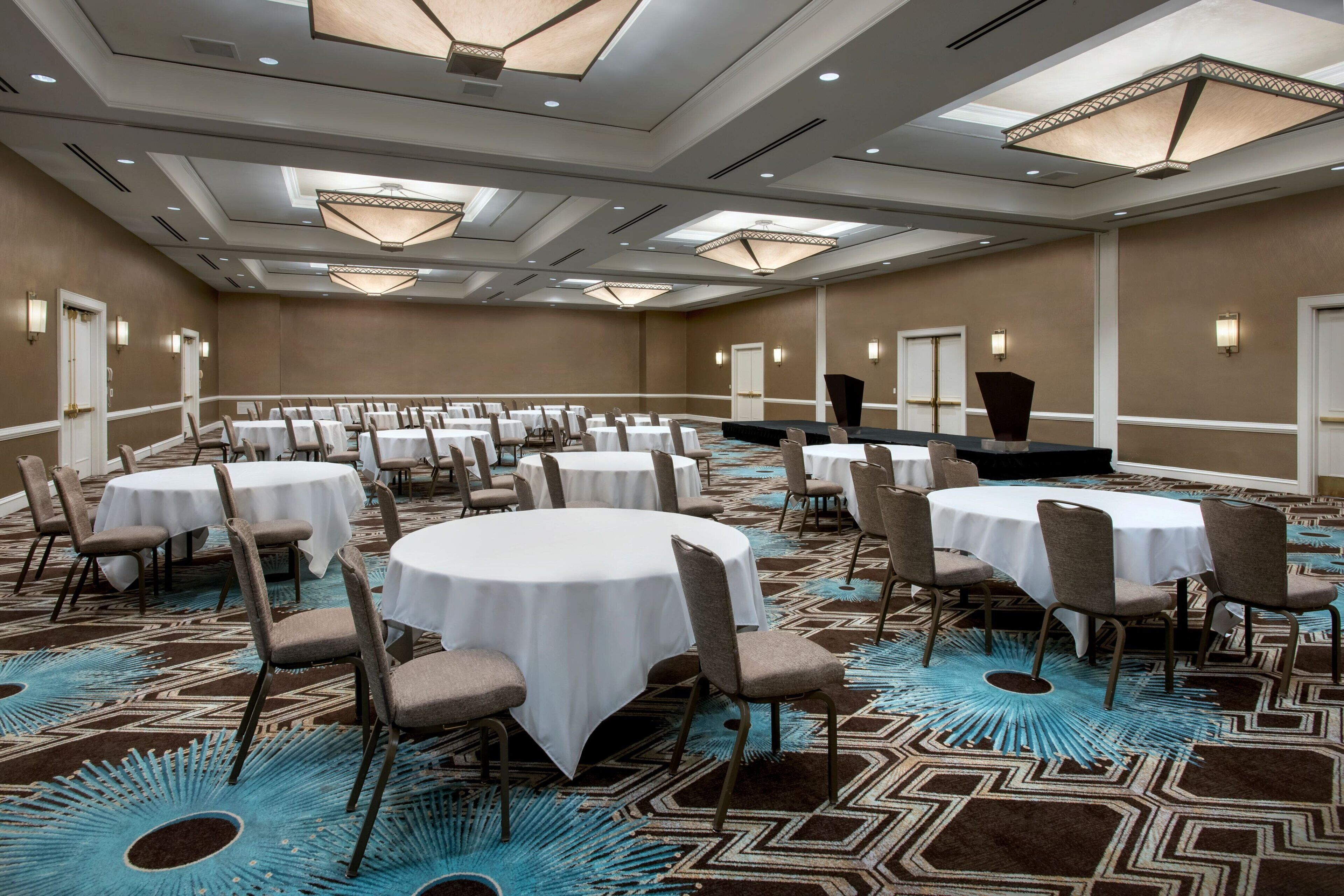 A large conference room with multiple round tables, each surrounded by chairs, and a stage at the front.