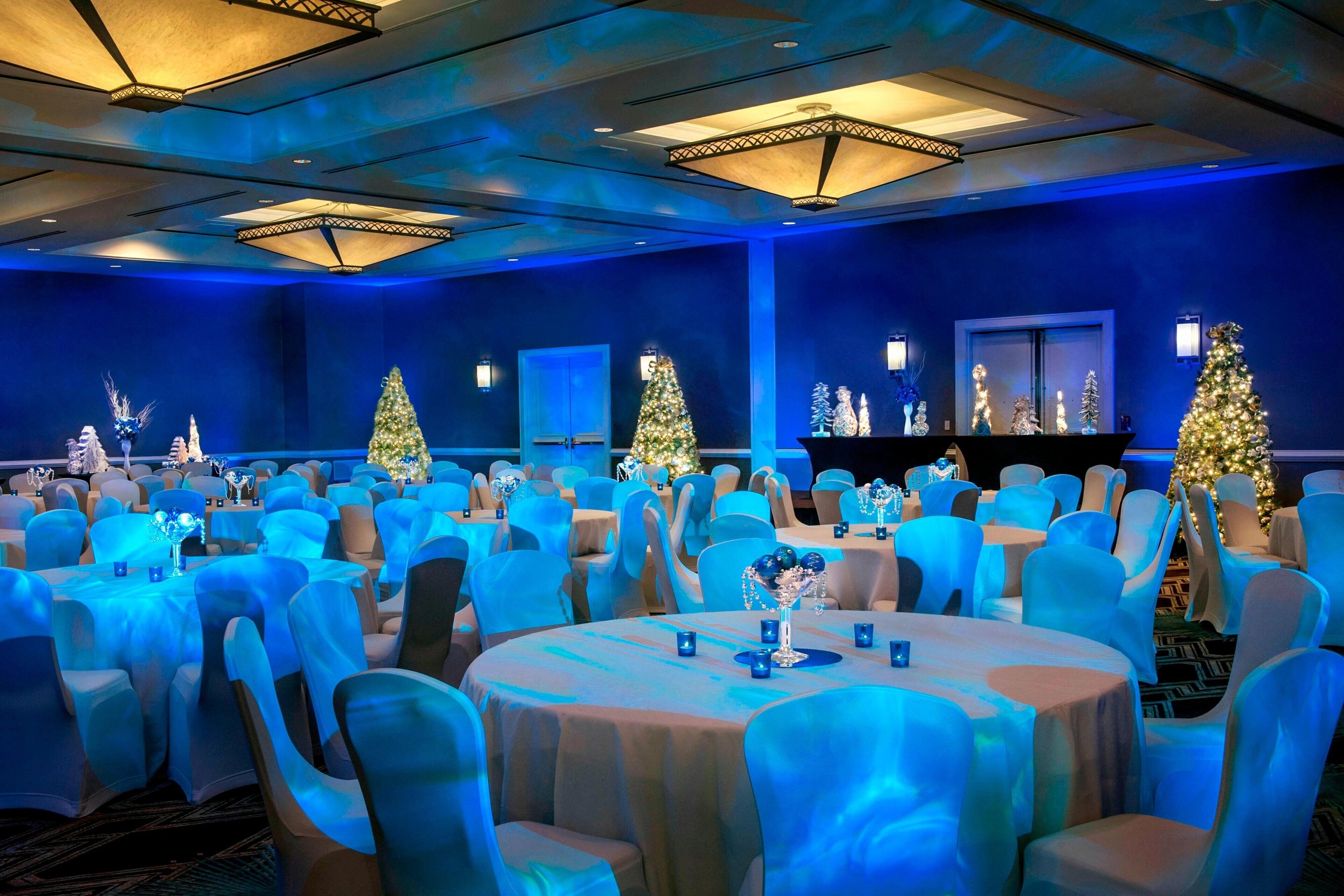 A banquet hall with round tables and blue chairs, decorated with Christmas trees and lights.