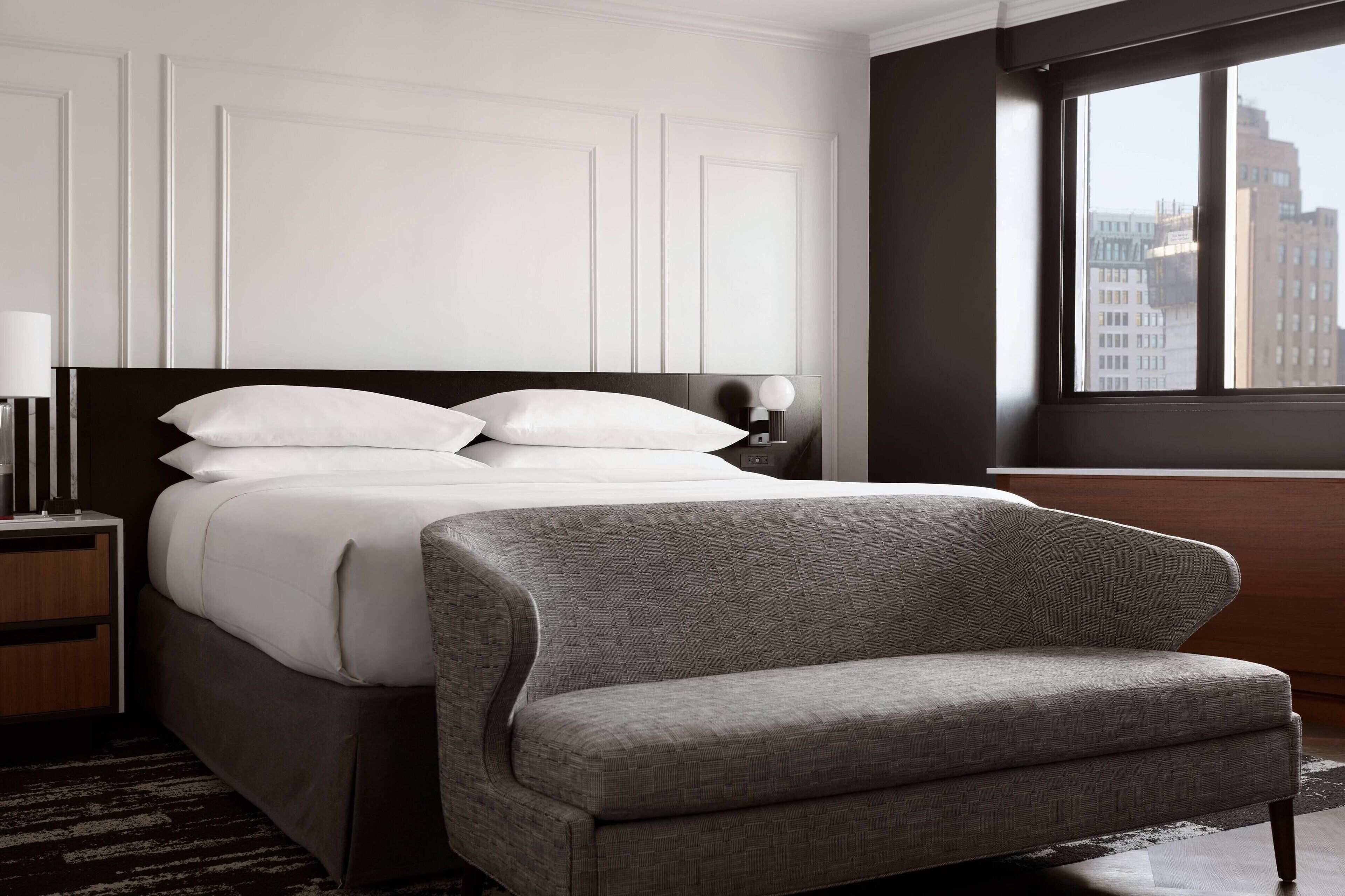 A modern hotel room with a large bed, a grey armchair, a nightstand with a lamp, and a window offering a city view.