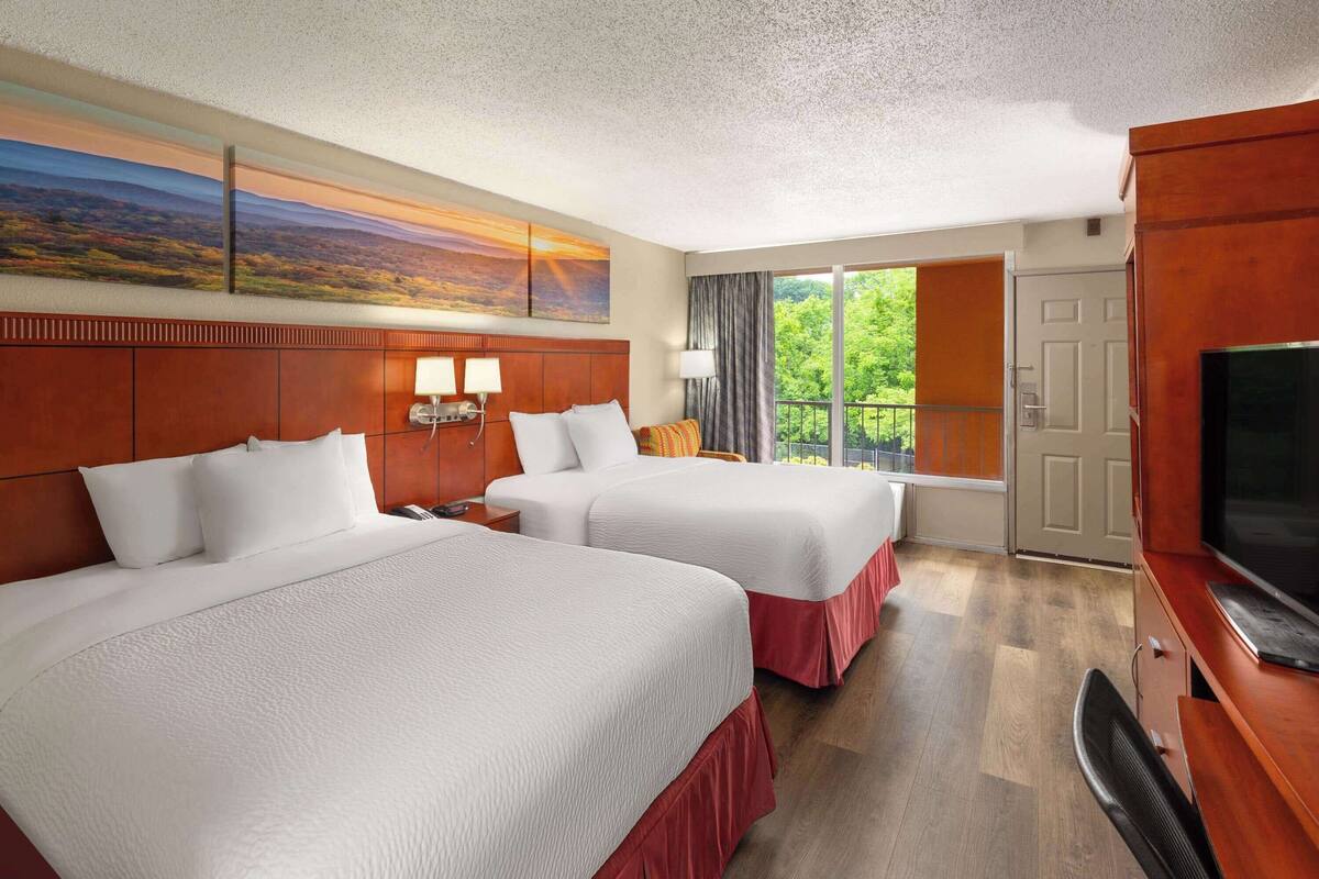 A hotel room with two beds, a TV, a large window with a view, and a painting on the wall.