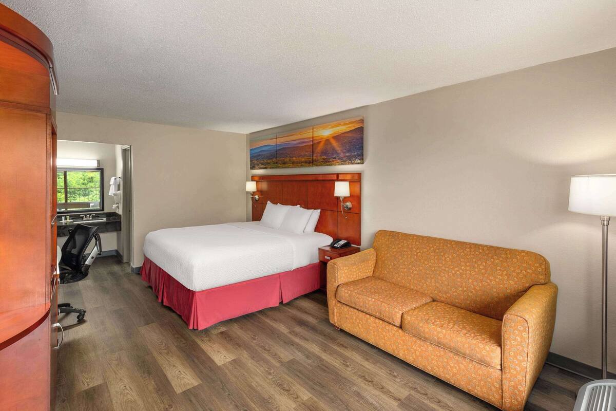 A hotel room with a bed, a sofa, a desk with a chair, a lamp, and a painting on the wall.