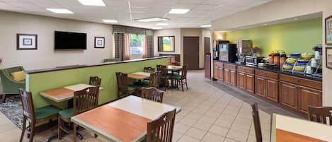 A hotel dining area with booths and tables, a kitchen counter with food service equipment, and a television mounted on the wall.