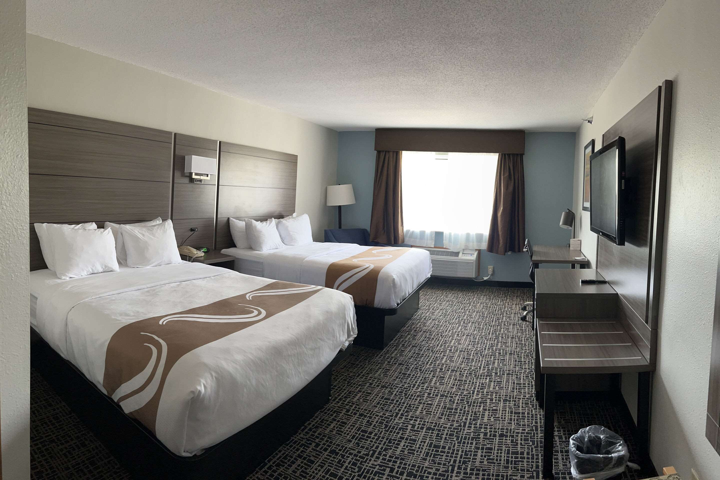A hotel room with two beds, a desk, a TV, and a window with curtains.