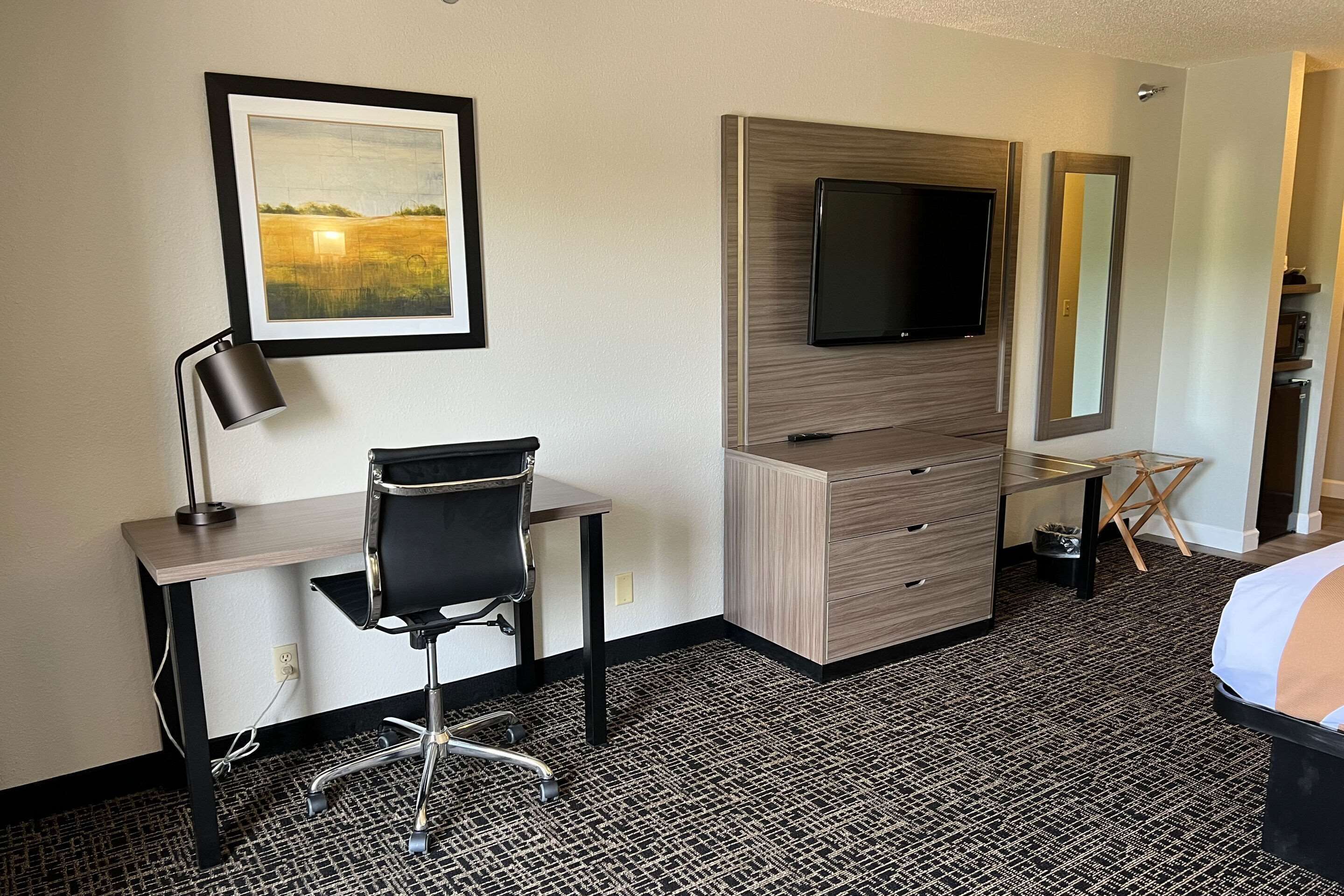 A hotel room with a desk, chair, TV, dresser, and a bed.